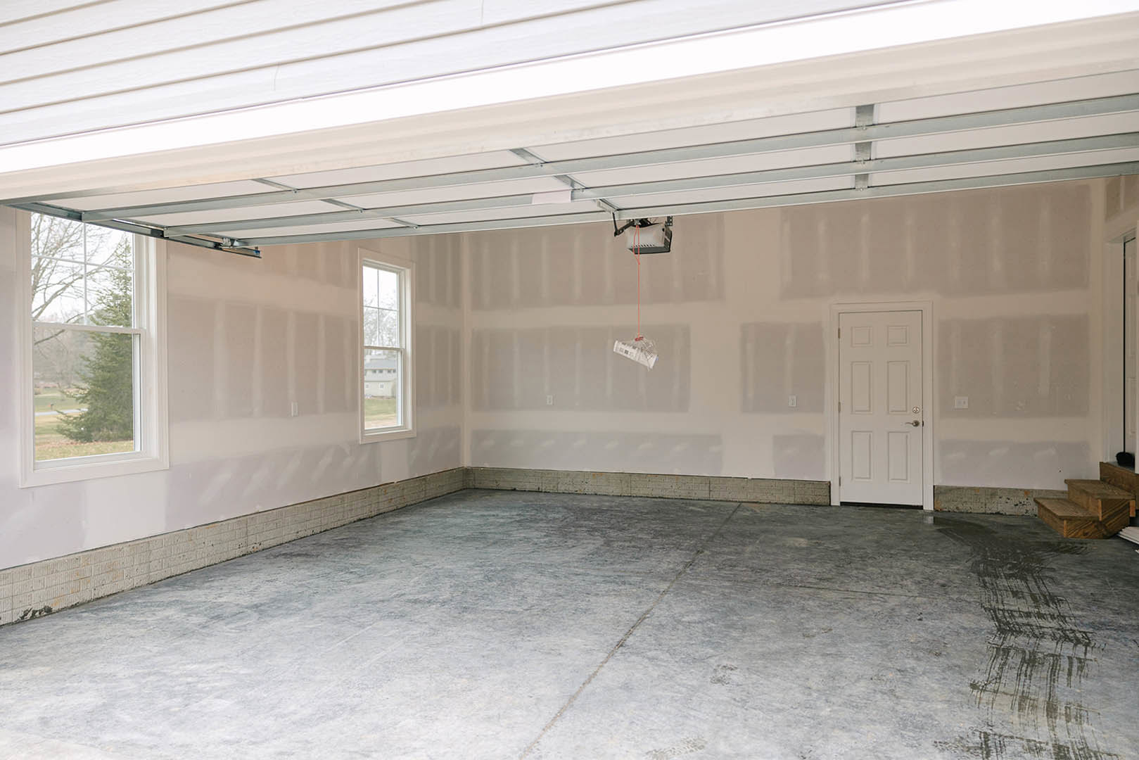 Garage interior with concrete floor, white walls, white garage door featuring a silver handle, ceiling detail, window overlooking trees, and a framed painting displayed on the