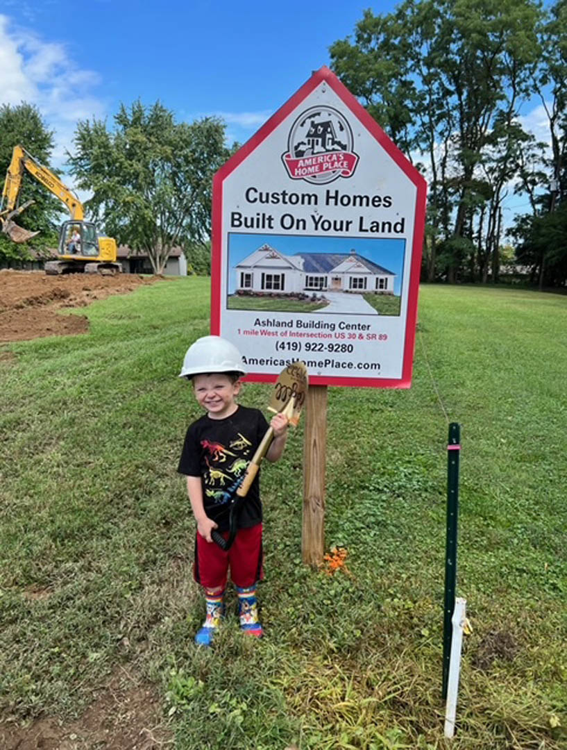 Young boy in jeans and t-shirt holding a shovel on grassy lawn, standing before a white sign with text, with trees and clouds in the background