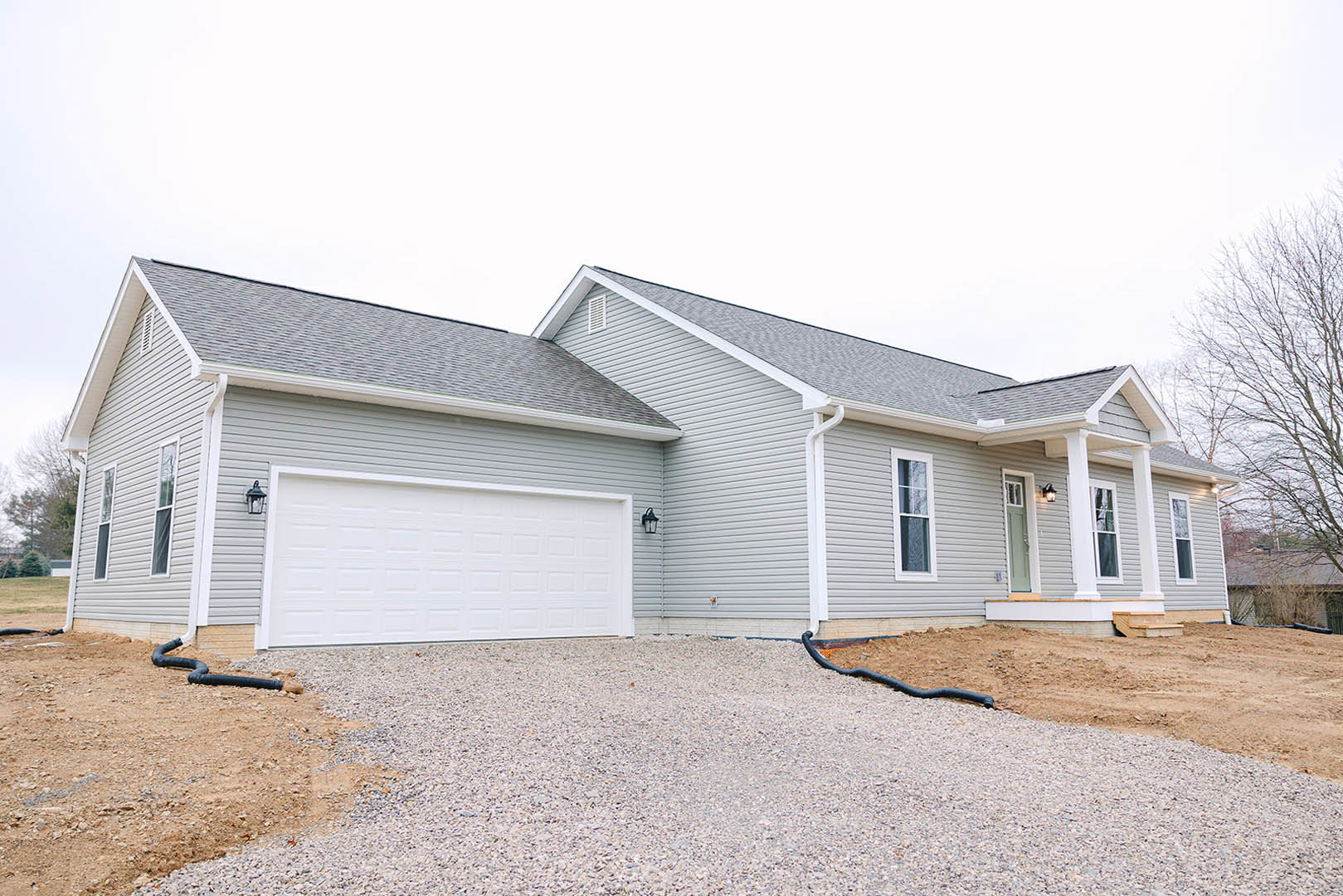 White garage door beneath a grey roof, white-framed window, gravel driveway with a dog standing near a black pipe, surrounded by siding and trees.