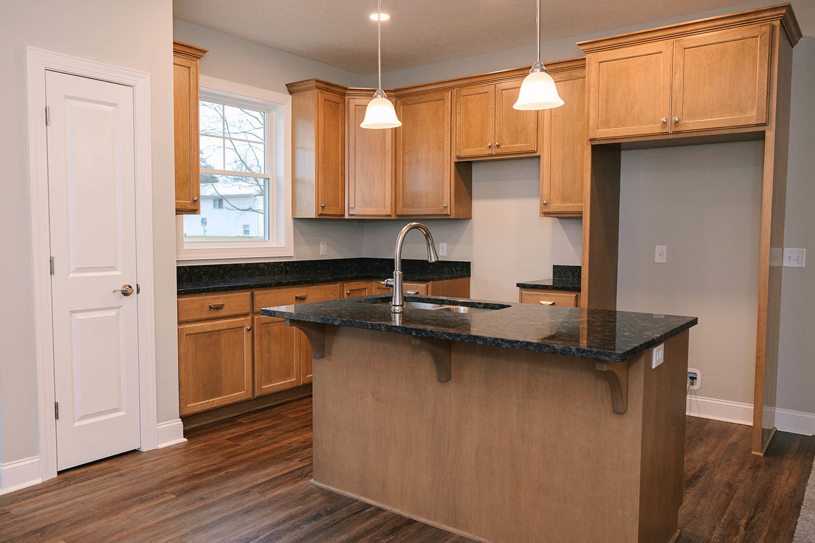 Kitchen with black stone countertop, wooden cabinets, island with built-in sink and modern faucet, white door, recessed lighting, and light switch on neutral wall