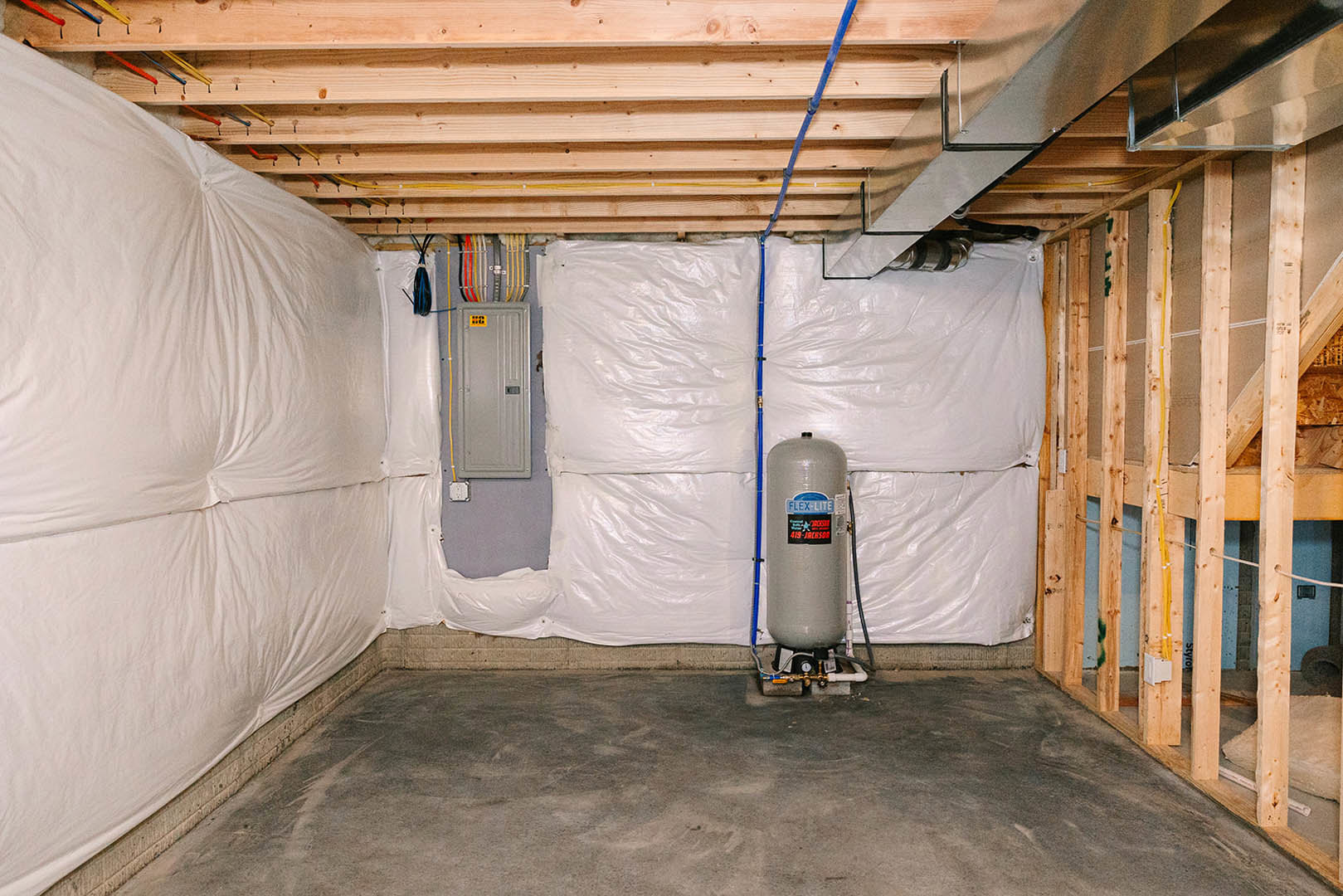 Basement room with exposed concrete floor, large grey cylindrical tank, white door with black handle, wood panel, and visible ceiling beams