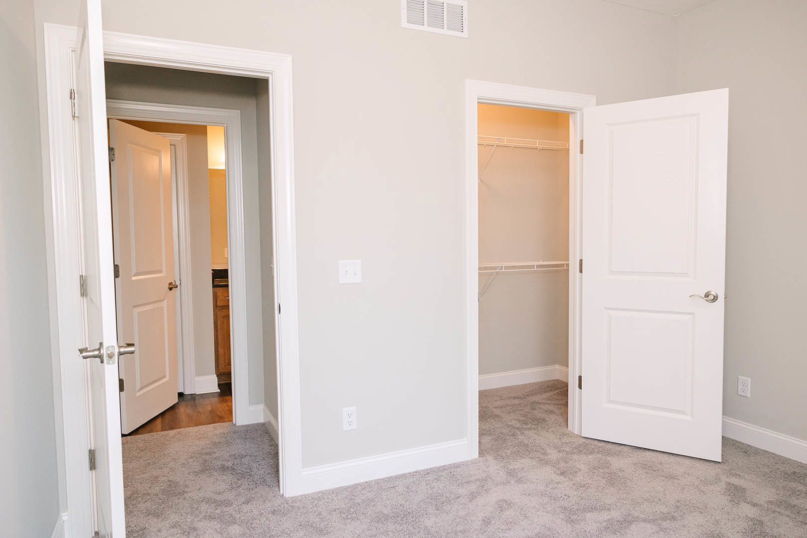 Bedroom with white paneled doors, built-in closets, silver door handle, white wall outlet with black plugs, white baseboard, and wall vent on light-colored plaster walls