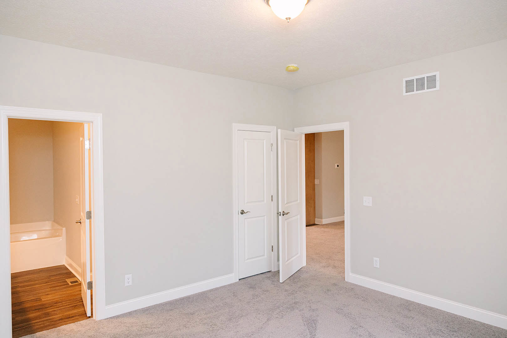 White walls and doors, wooden floor with vent, ceiling light fixture, white door with silver handle, wall vent, bathroom featuring white bathtub