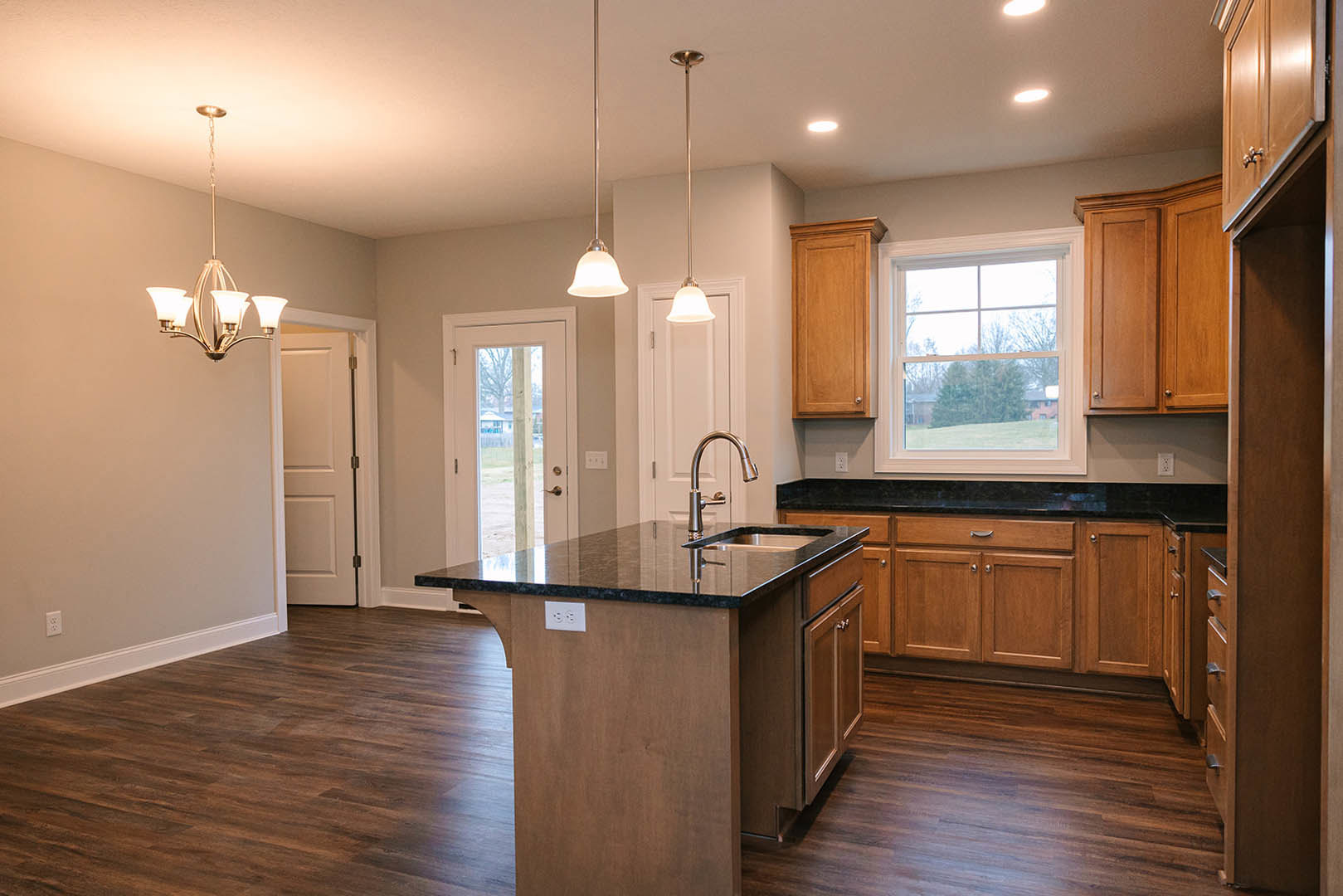 Open kitchen with wood flooring, central island featuring built-in sink, white cabinetry, large window overlooking trees, wooden door with light frame, glass-paneled door, and