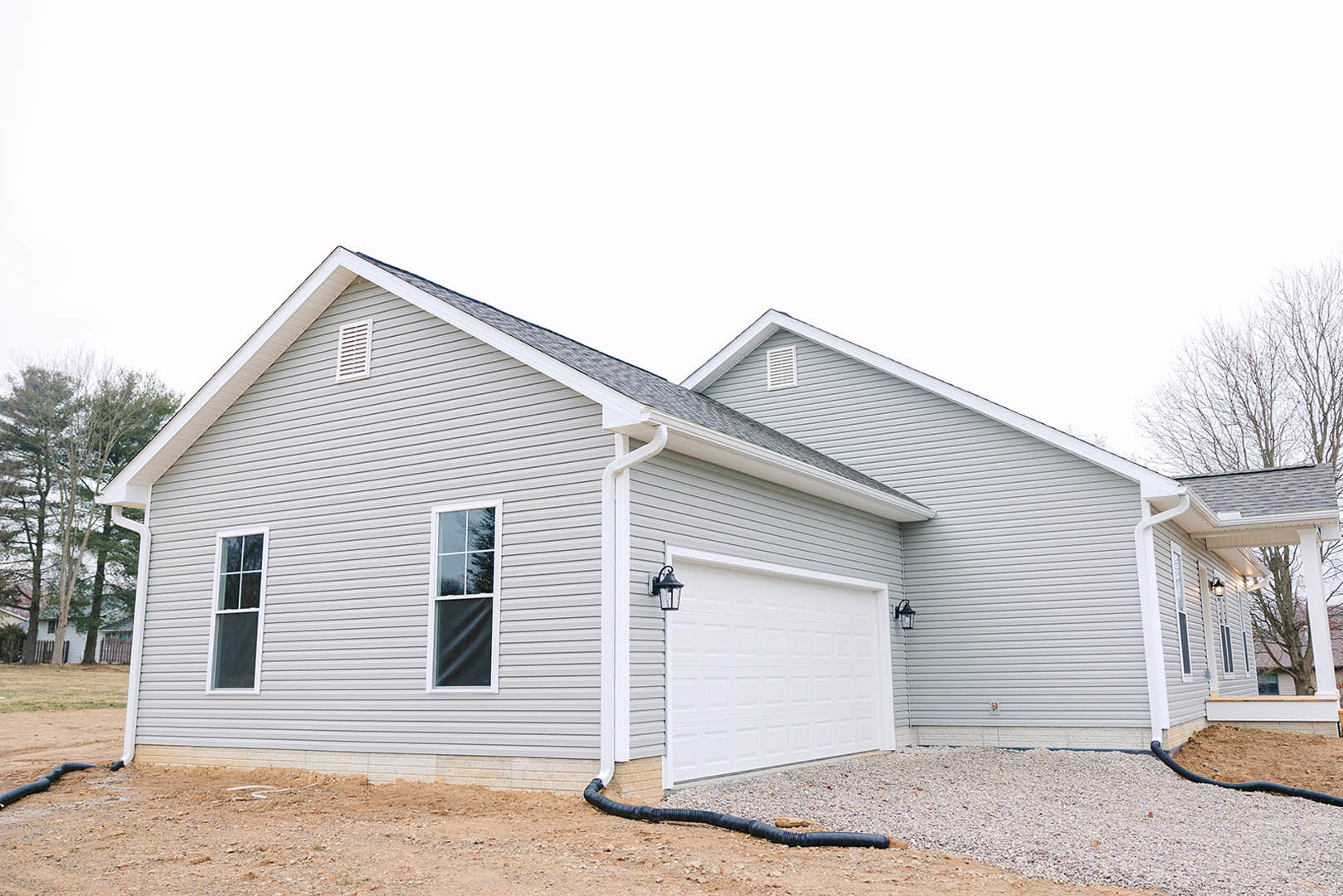 Two-story home with light-colored siding, white-framed windows, attached single-car garage with white door and exterior lamp, white vent above garage, black drainage pipe along