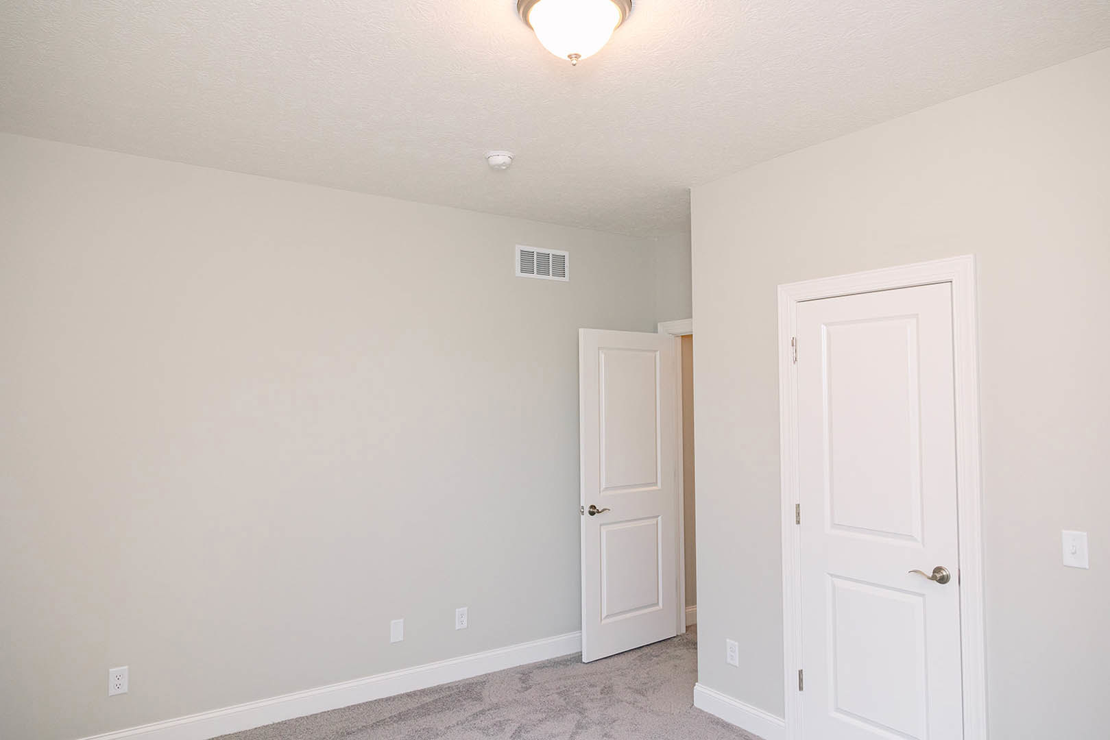 Room with white walls, two white doors with silver handles, ceiling-mounted light fixture, wall vent, and light-colored flooring.