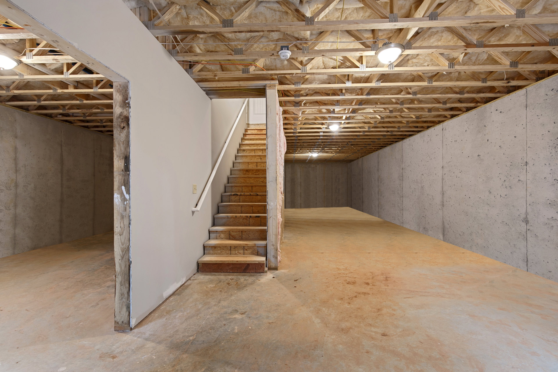 Wood staircase with open risers and metal railing, light fixture hanging from plaster ceiling, concrete floor, exposed beams, and composite material walls in modern basement