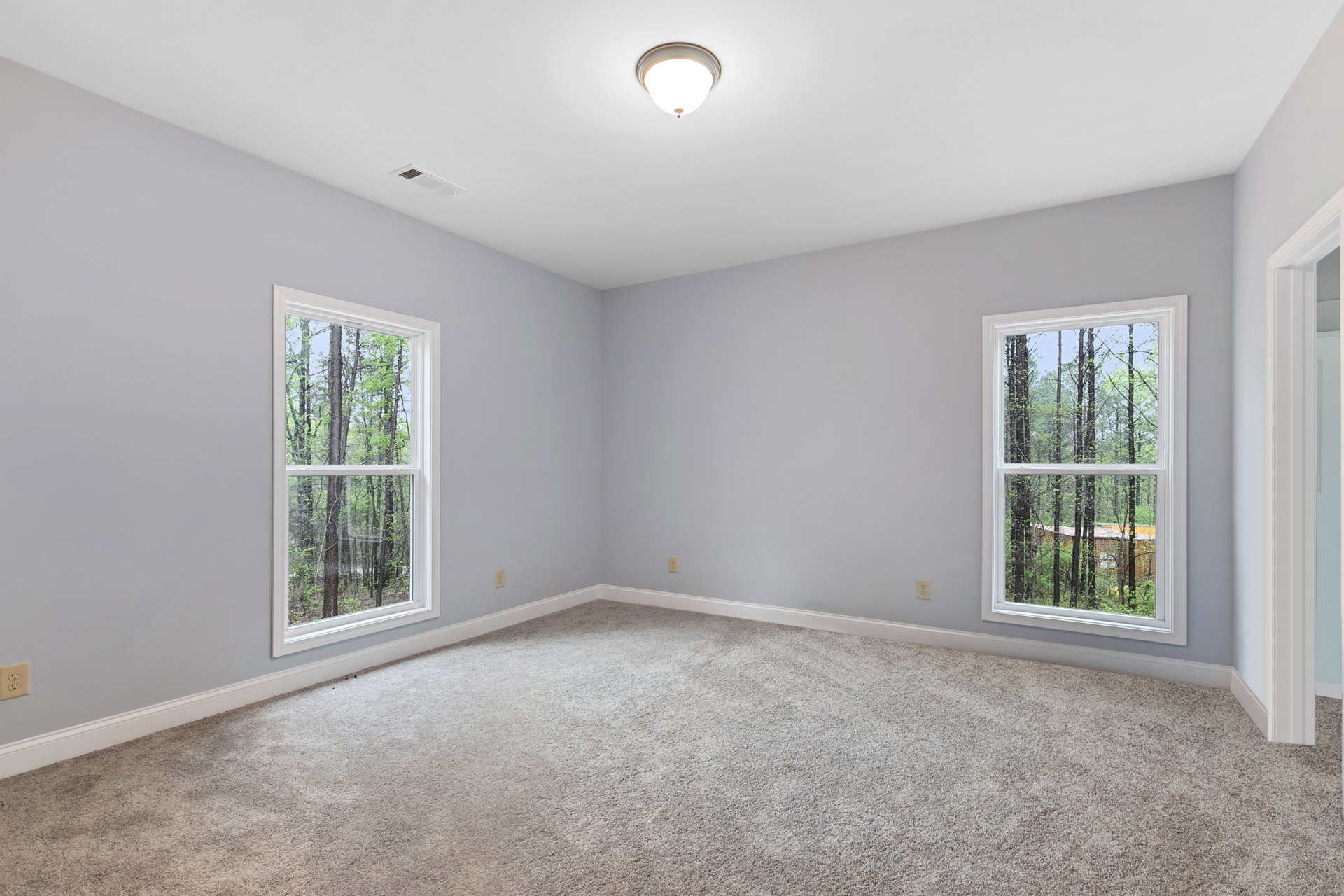 Bright carpeted room with large windows, white walls, ceiling light fixture, and tree views outside