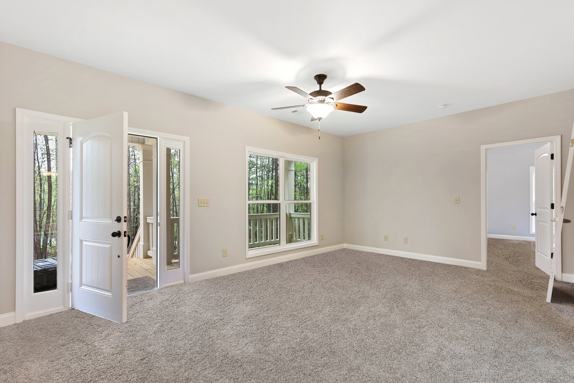 White ceiling fan with integrated light fixture centered above hardwood floor; white walls, single window with view of green trees, and paneled door with black hardware.