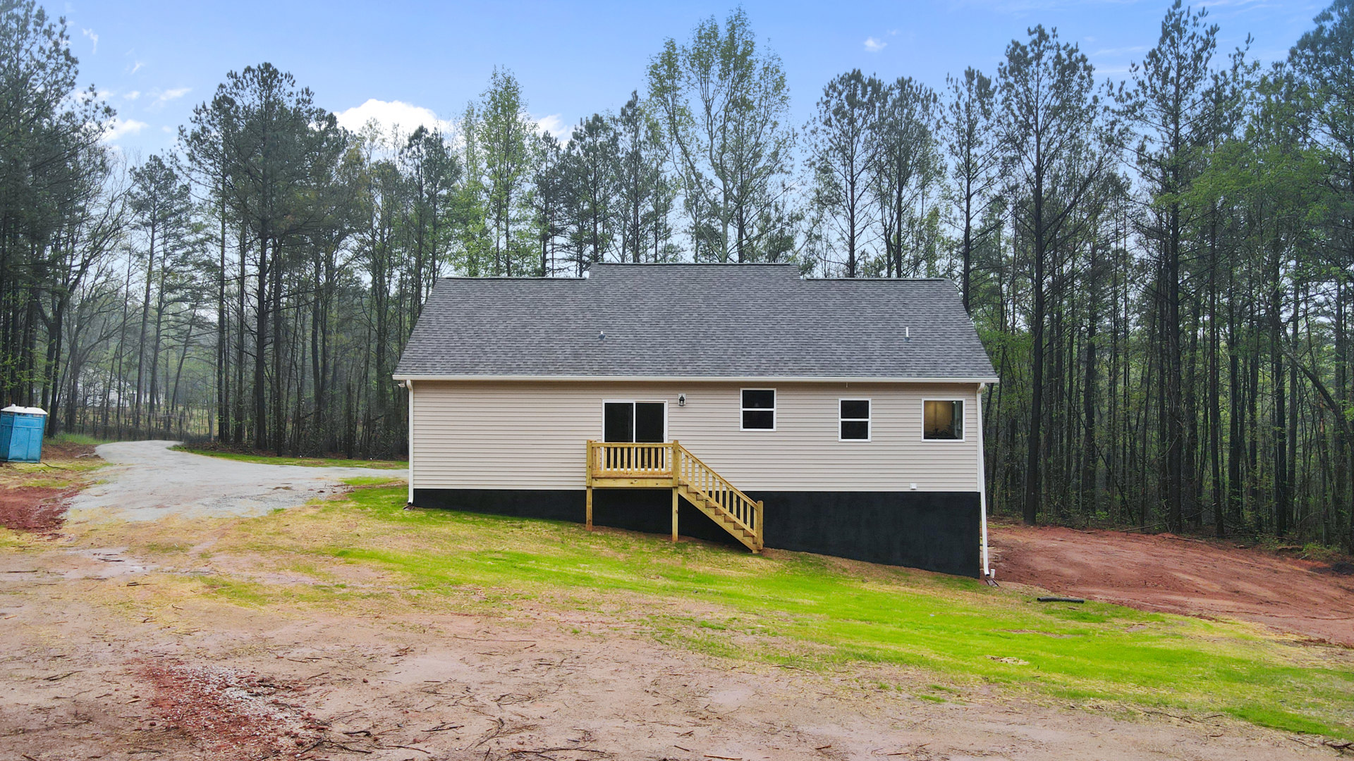 Two-story house with wooden staircase and deck, white-framed windows, blue portable toilet on lawn, surrounded by grass and trees under partly cloudy sky