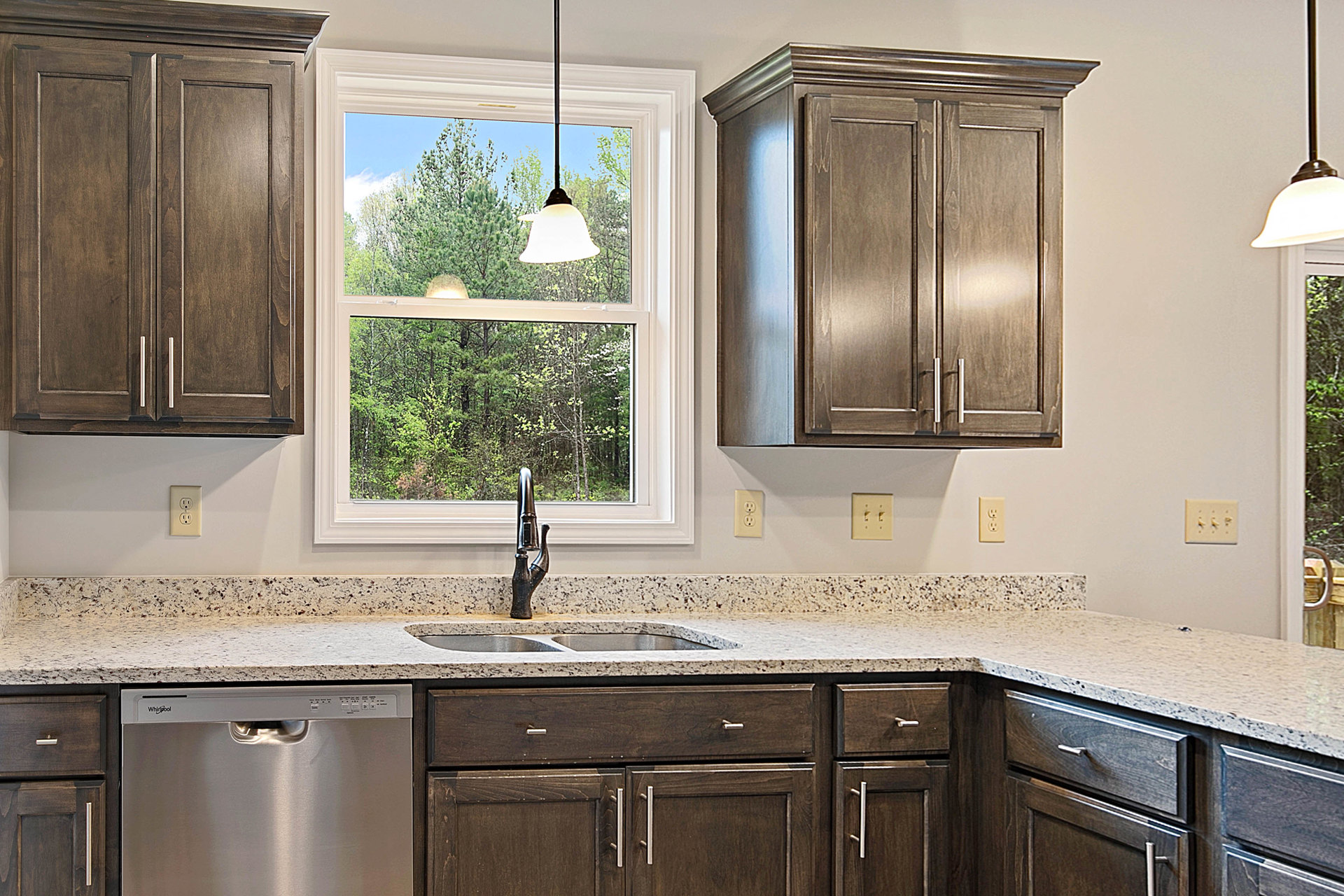 Modern kitchen featuring white cabinets with metal handles, stainless steel sink and faucet, built-in dishwasher, light switch on a neutral wall, large window with blue sky, wood