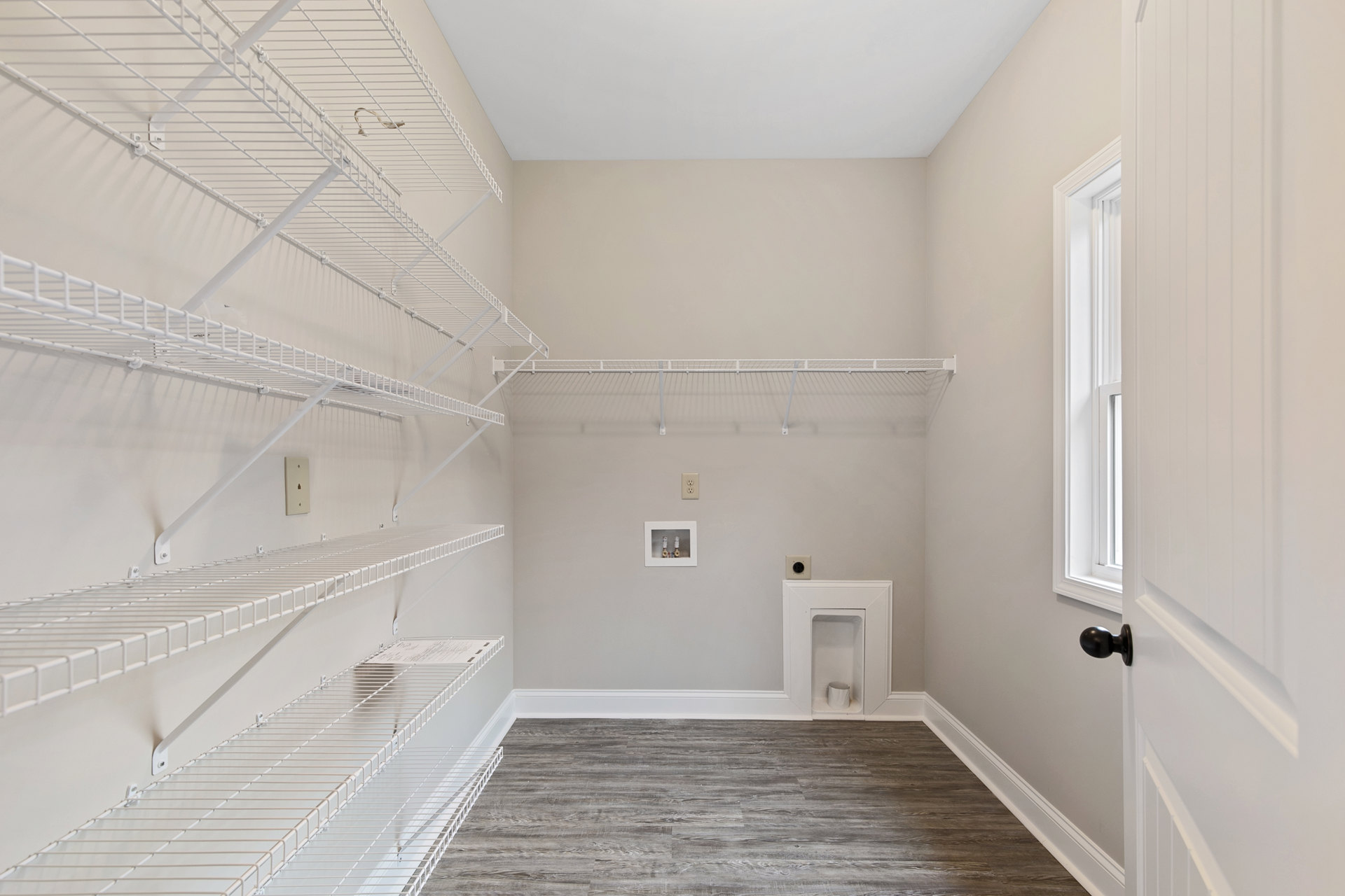 Empty room with built-in white shelves, light wood flooring, white walls, black door knob, window, and small wall opening with exposed pipes