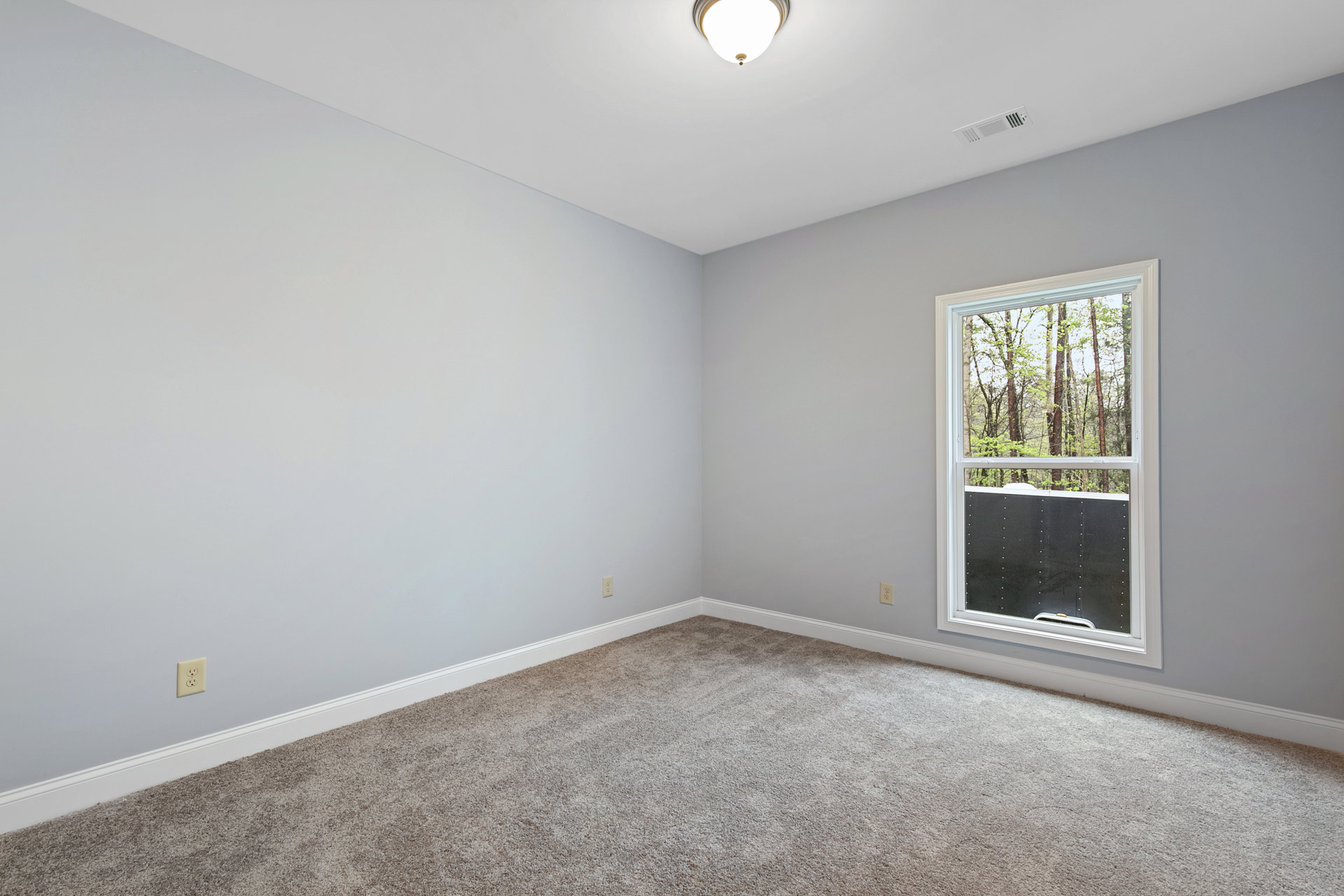 Neutral-toned carpeted room with white walls, large window overlooking trees, circular ceiling light, and black window trim