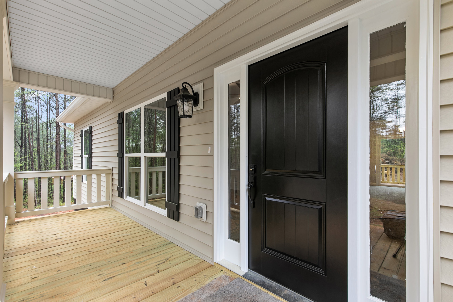 Black front door with silver handle, wooden porch deck, white railing, wall-mounted lamp, plastic wall cover, exterior house window