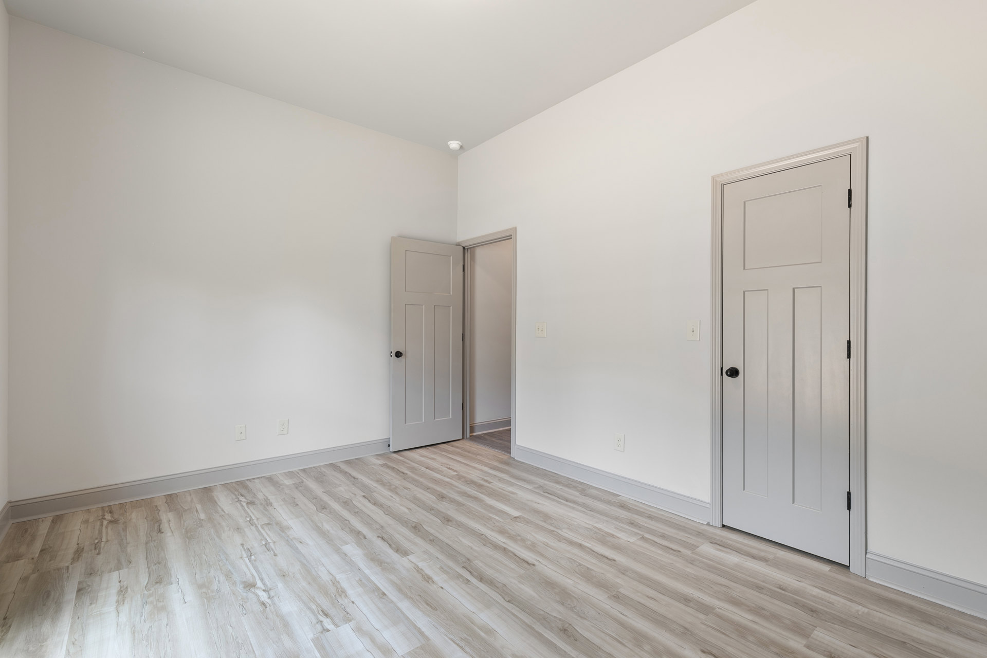 Room with light wood flooring, two white doors featuring black hardware, white walls, and smooth plaster ceiling