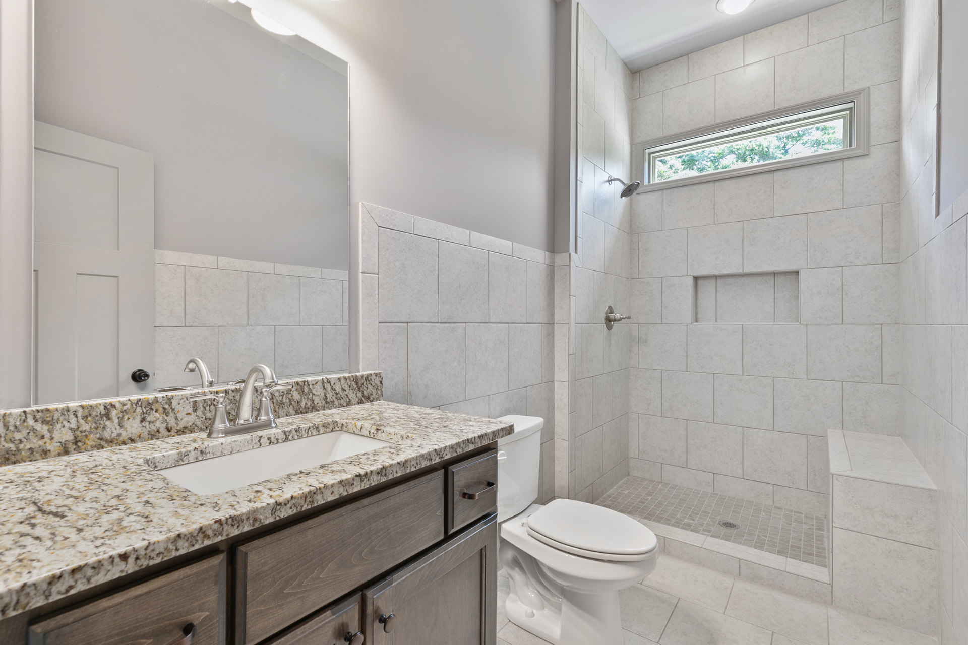 Modern bathroom with white toilet, rectangular sink on wood vanity, light gray tile walls, and frosted window above countertop