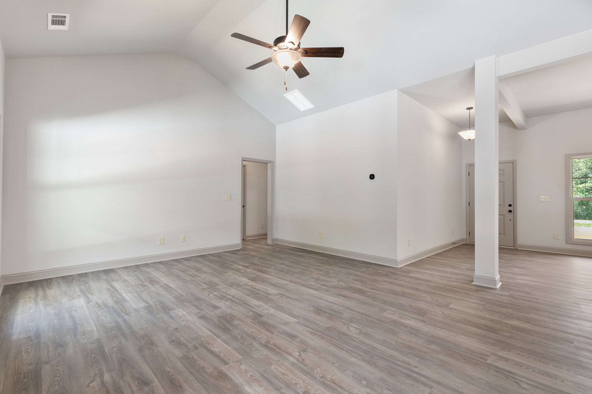 Ceiling fan with light fixture, wood flooring, white walls, window with white frame, white door with black handle, and wall vent in a custom home interior.