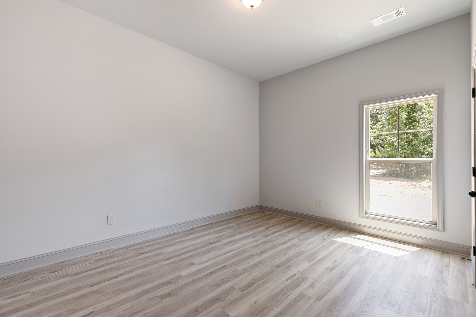 Sunlit room with wide window overlooking trees, wood plank flooring, white plaster walls, ceiling vent, and modern light fixture