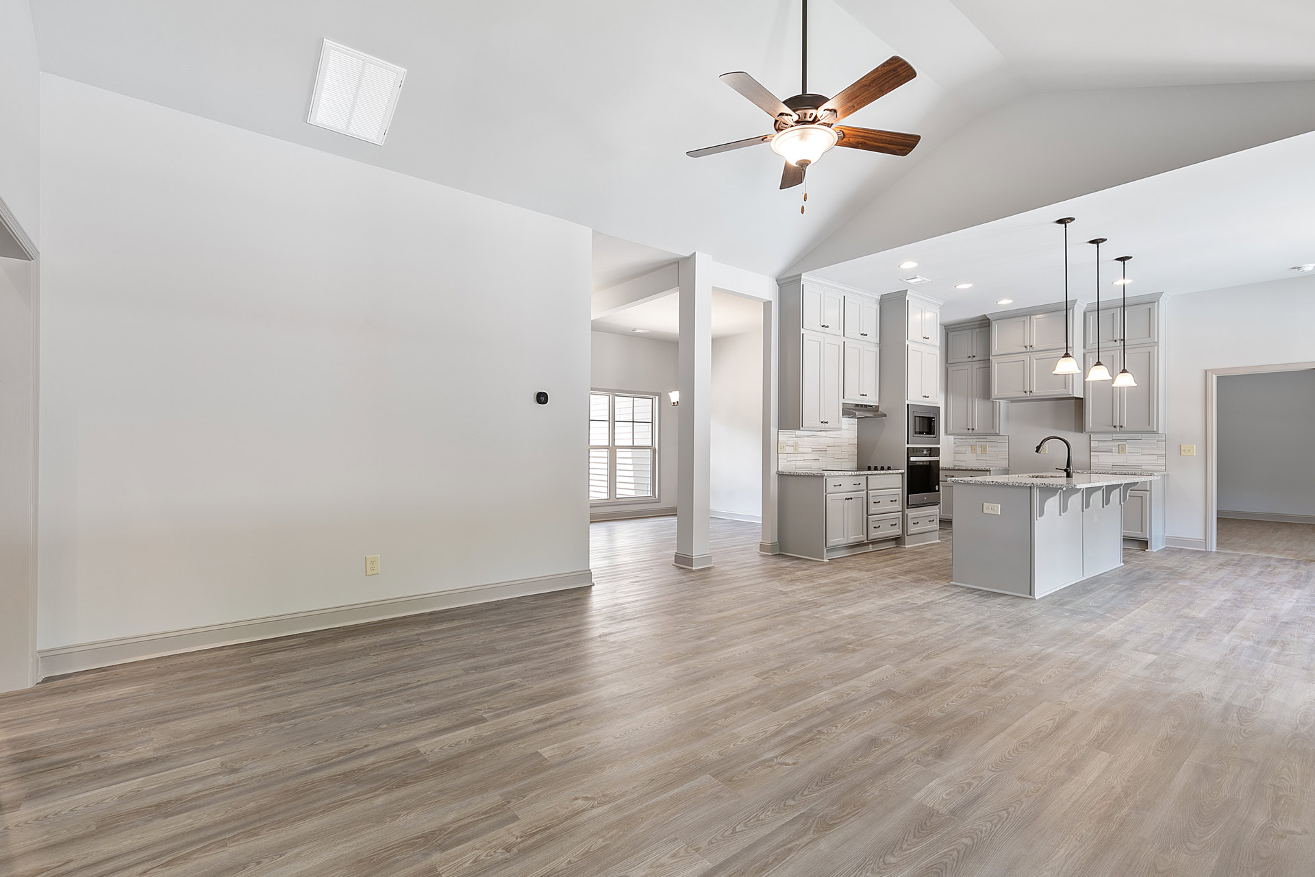 Open-concept kitchen and living room featuring a wood floor, kitchen island with sink, ceiling fan with light, white wall vent, plaster walls, and a close-up window.
