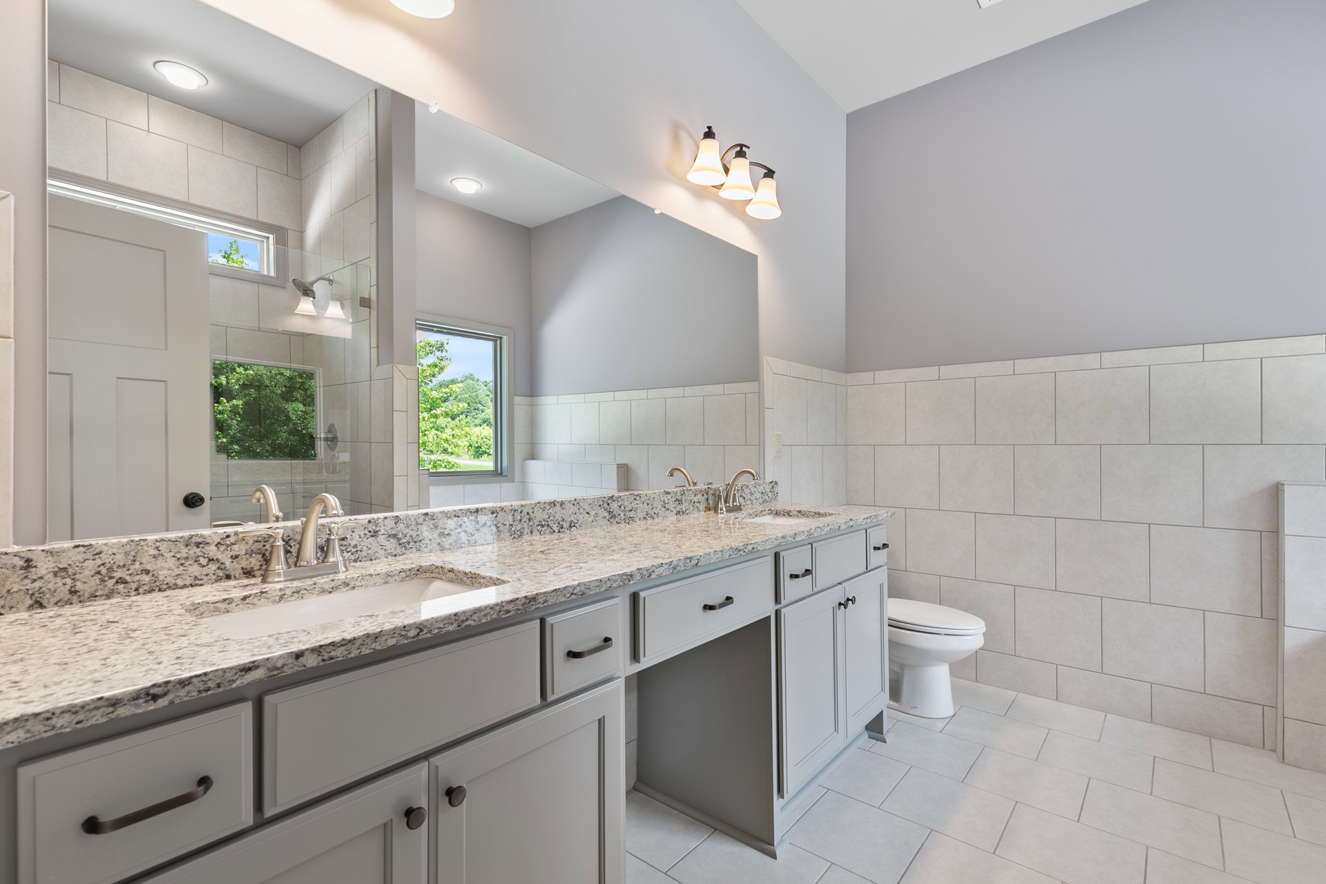 Bathroom with marble countertop, wide framed mirror above sink, modern faucet, tile backsplash, cabinetry with drawers, window overlooking trees, toilet partially visible
