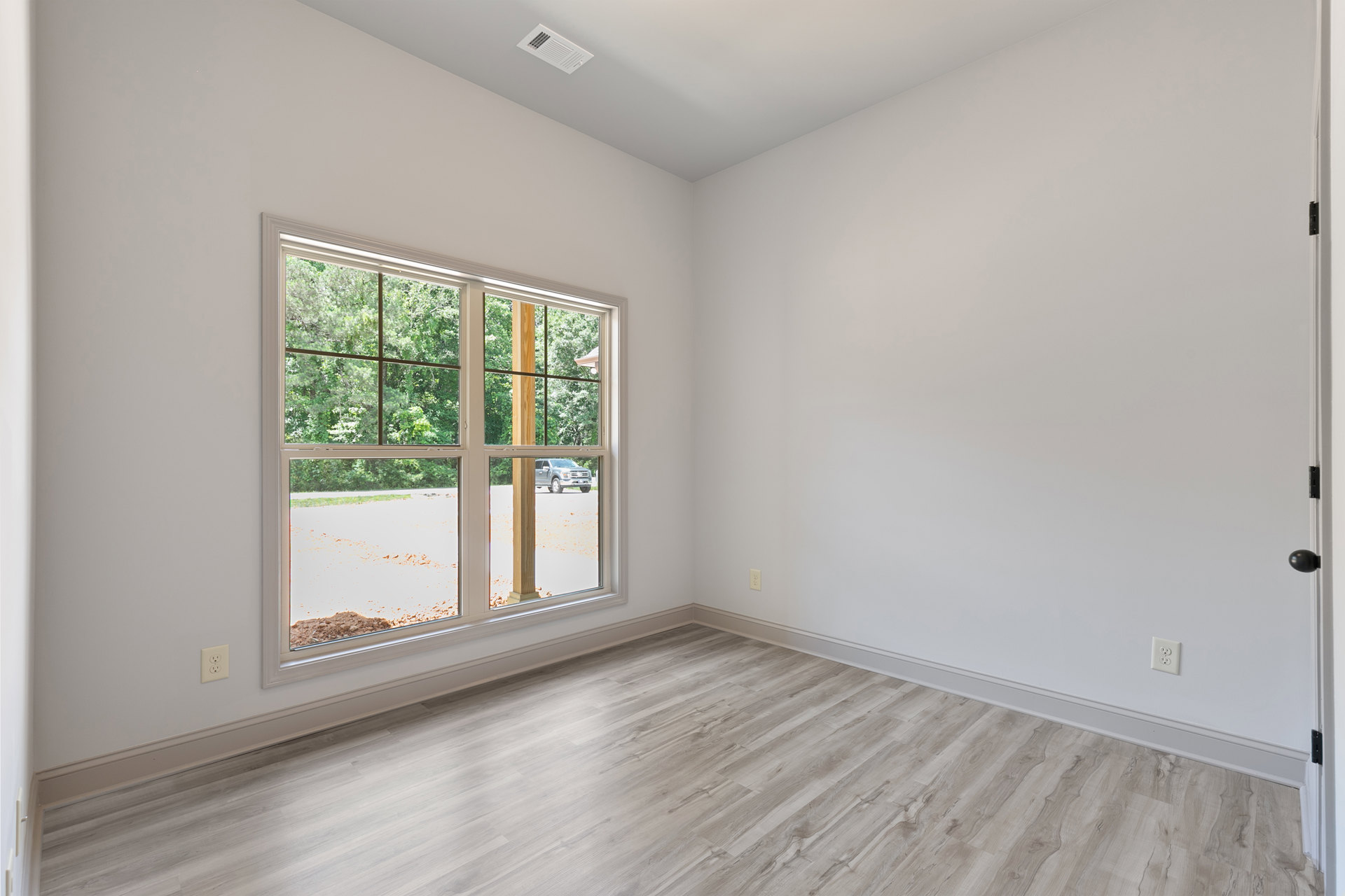 Sunlit room featuring a large window, light hardwood flooring, white plaster walls, and a ceiling vent.