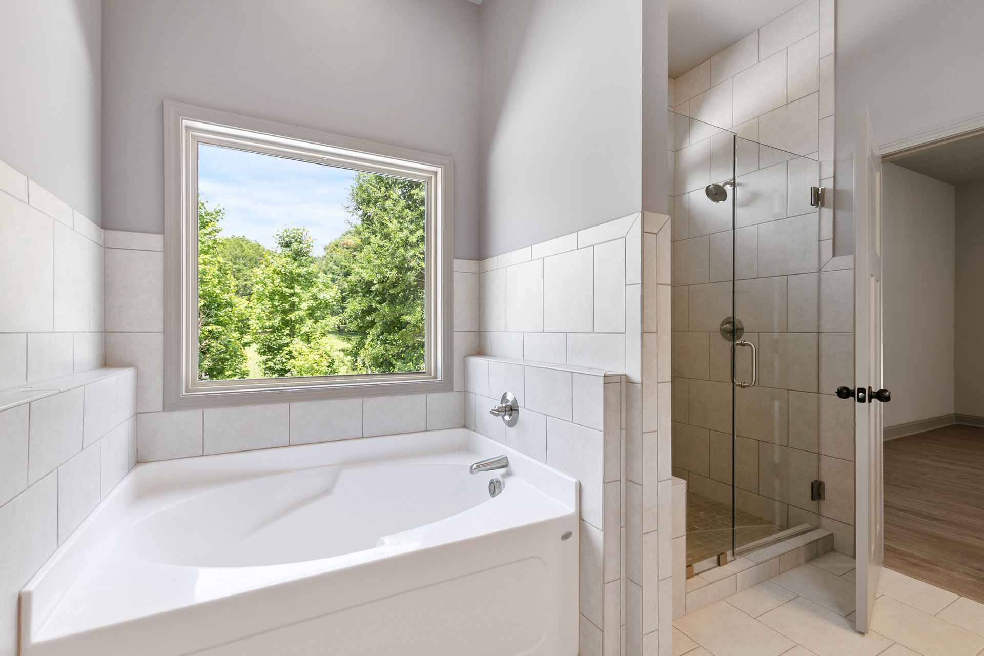 White freestanding bathtub beneath a large window with tree views, wall-mounted chrome faucet, light gray tile backsplash, natural wood flooring, brushed metal tub handle