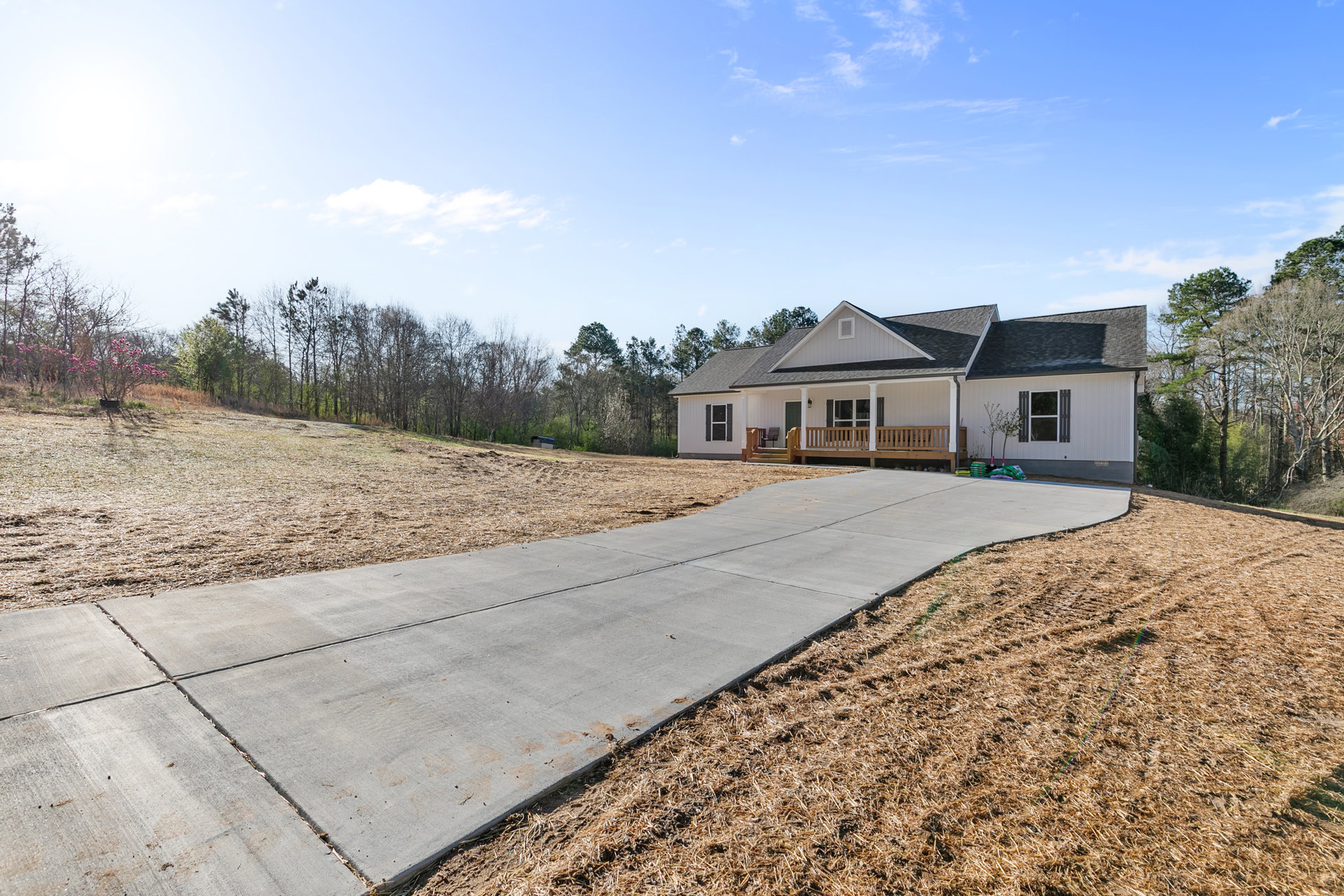 Concrete driveway leading to a single-story home with white-framed windows, wooden porch railing, and mature trees in the background