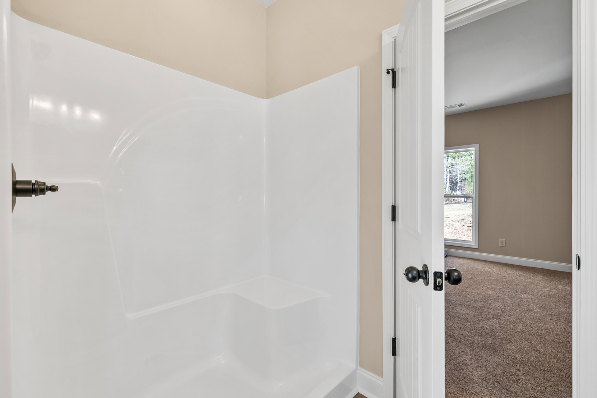 Freestanding white bathtub near open door, window overlooking trees and grass, black round door knob, plaster walls, modern bathroom fixtures