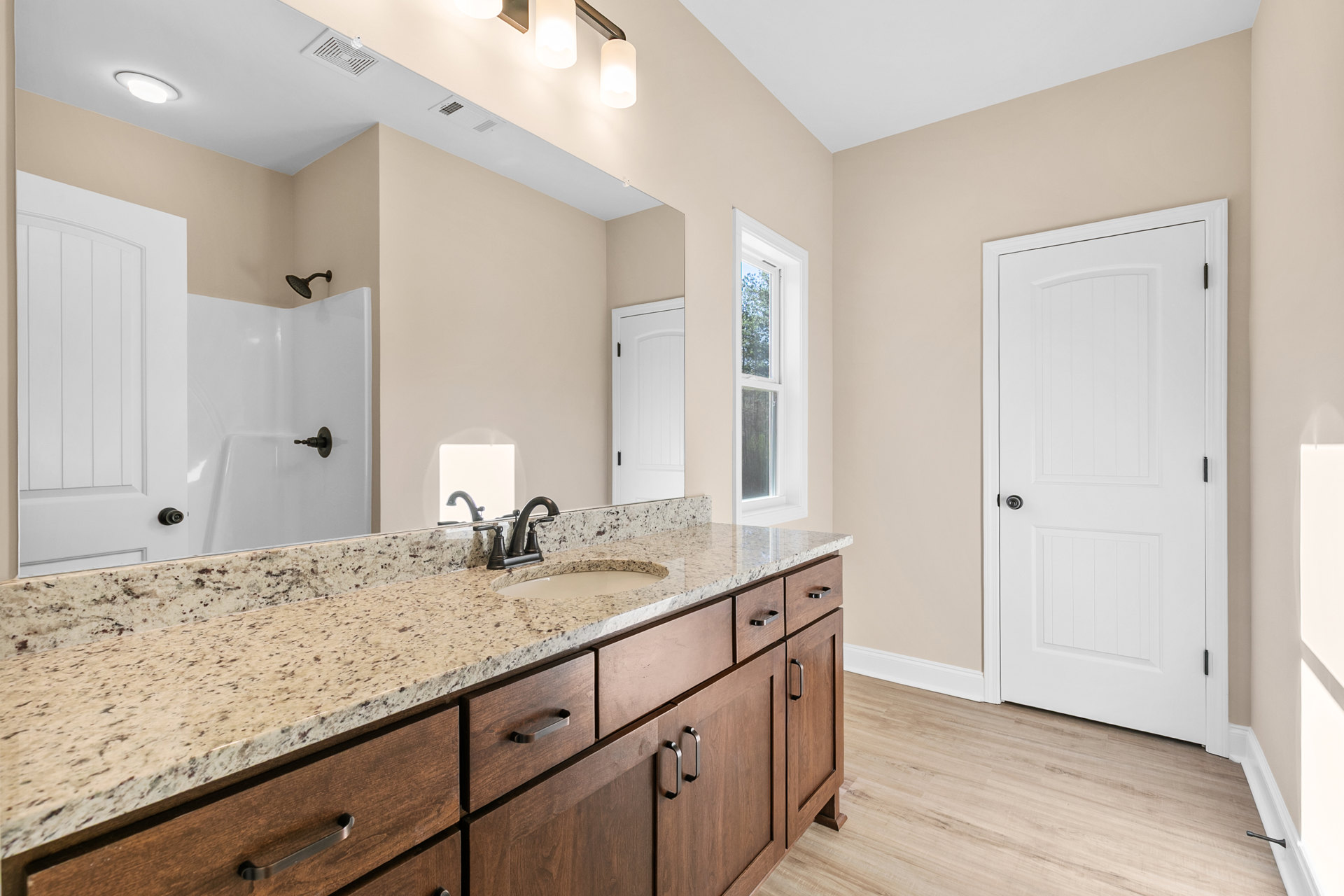 Bathroom featuring a marble countertop with undermount sink, large framed mirror, glass-enclosed shower, white door with black handle, recessed lighting, and window overlooking
