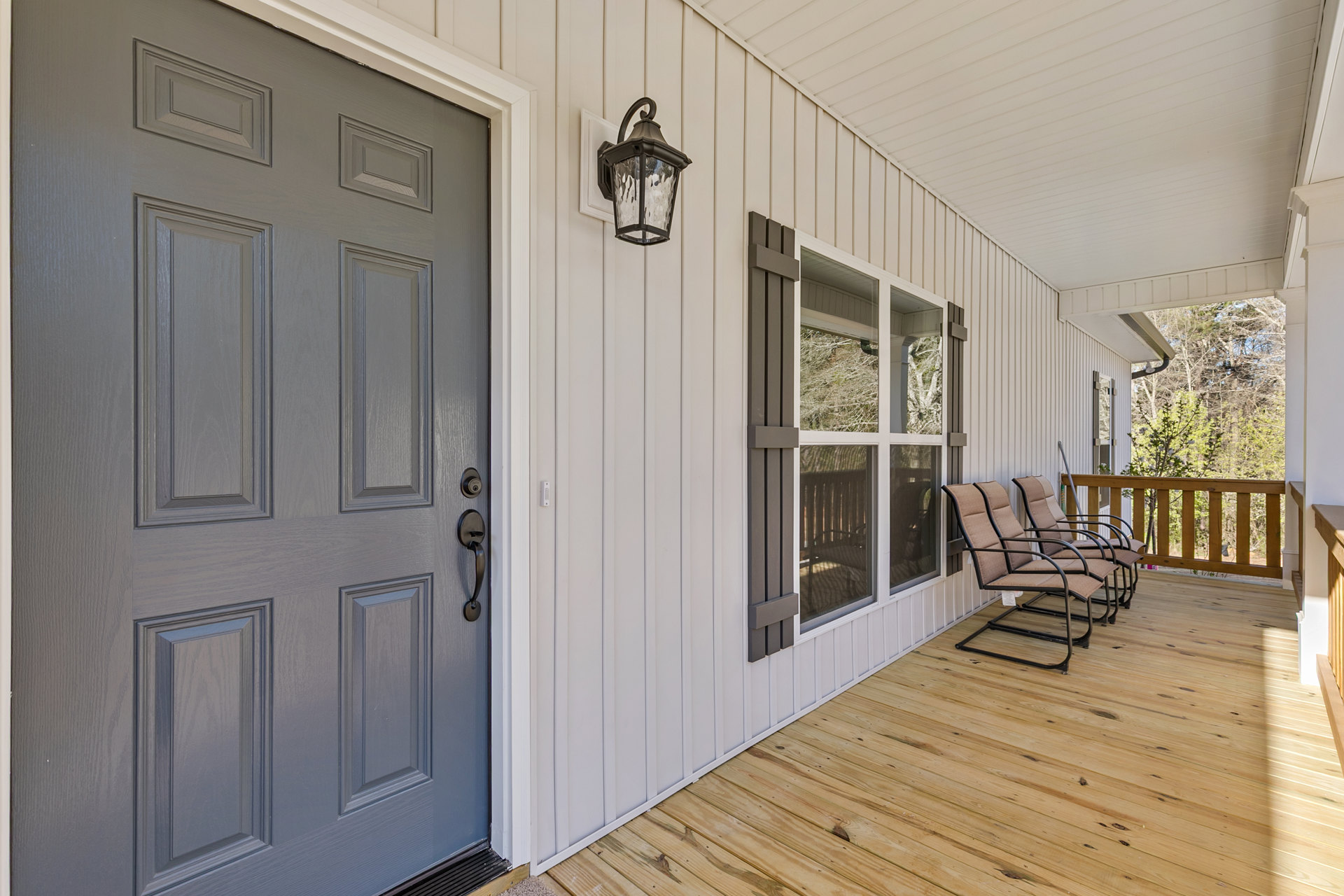 Wide front porch with wooden floorboards, white railing, several cushioned chairs, and a blue entry door with black hardware; leafy trees visible in the background.