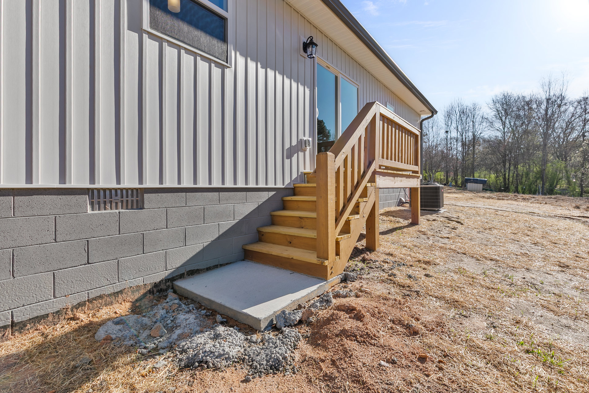 Wooden exterior stairs leading to a concrete porch, surrounded by trees and greenery, with a metal utility box and house roof visible in the background.