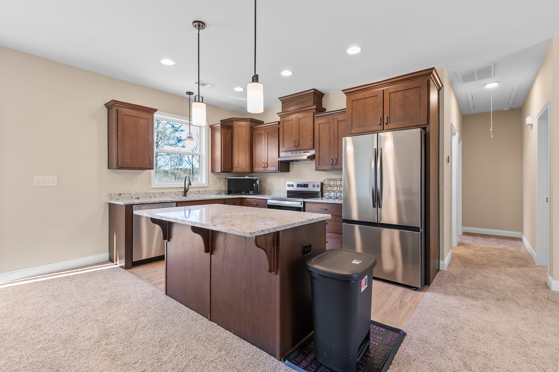 Kitchen with white granite island countertop, stainless steel refrigerator, black microwave, cabinetry, and black lidded trash can