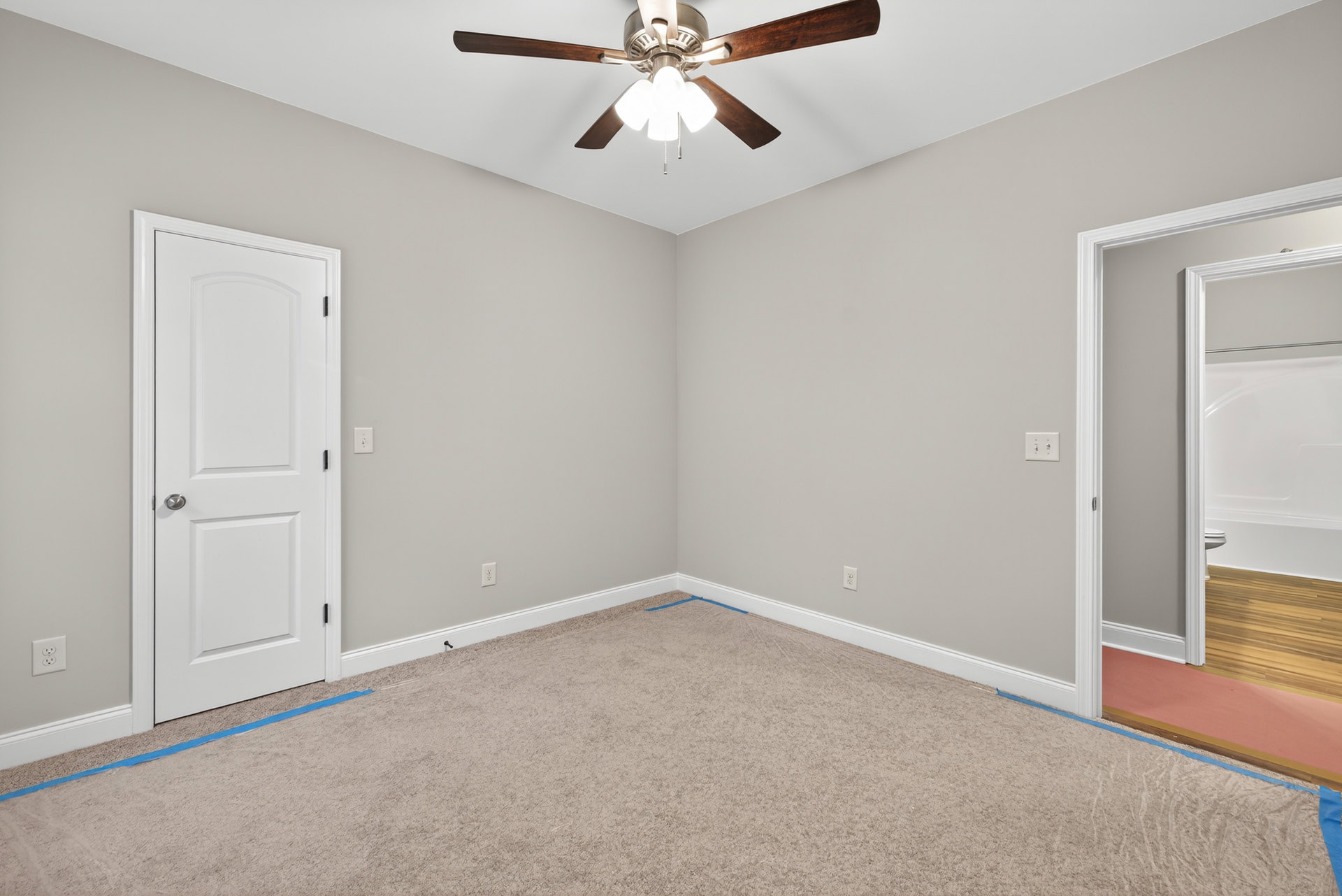 Ceiling fan with lights mounted above a room featuring a wood floor partially covered by a pink carpet, white door with silver handle, and blue tape on beige carpet.
