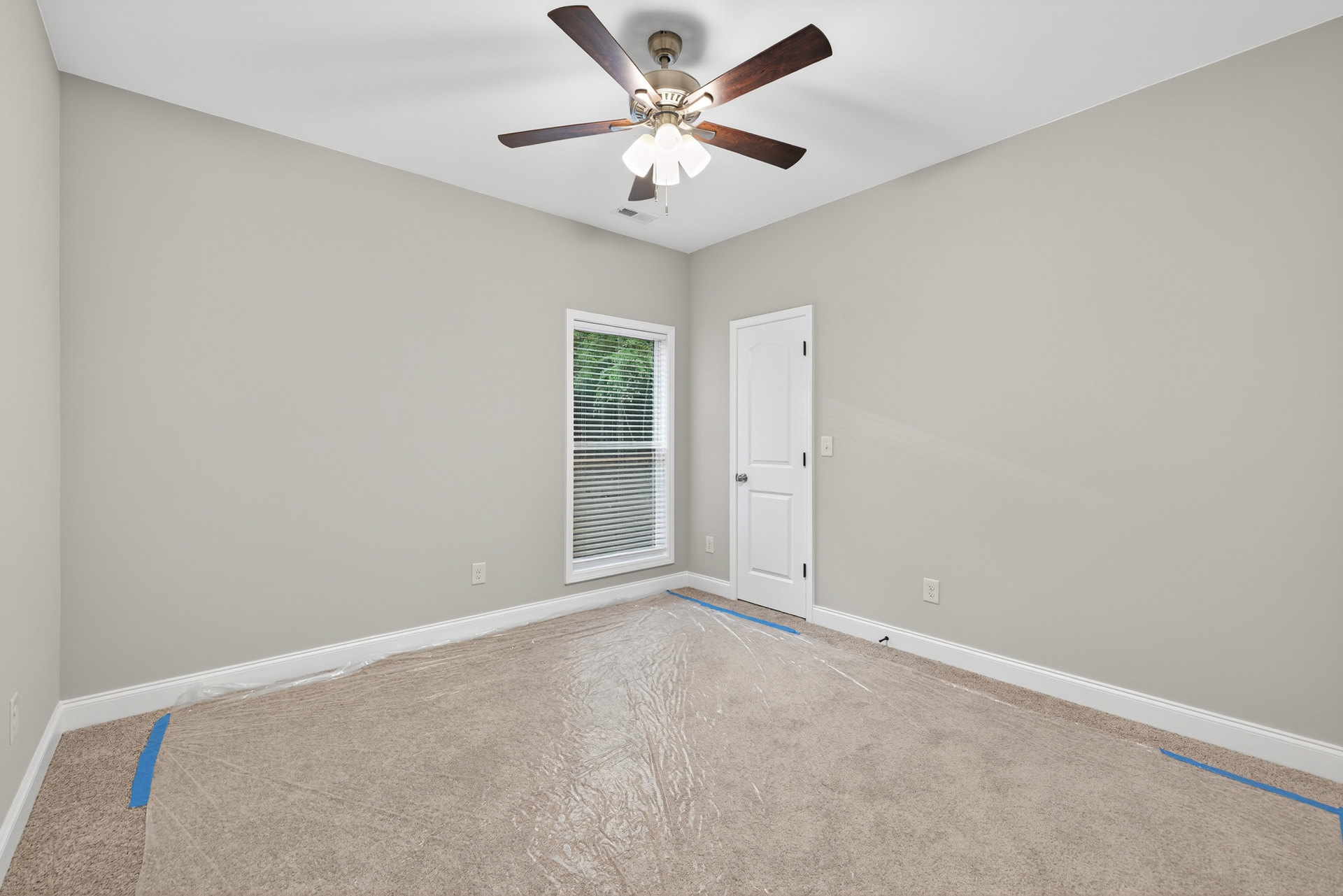 Ceiling fan with lights mounted above carpeted floor, white painted walls, window with blinds, white door with silver handle, blue strip on brown surface