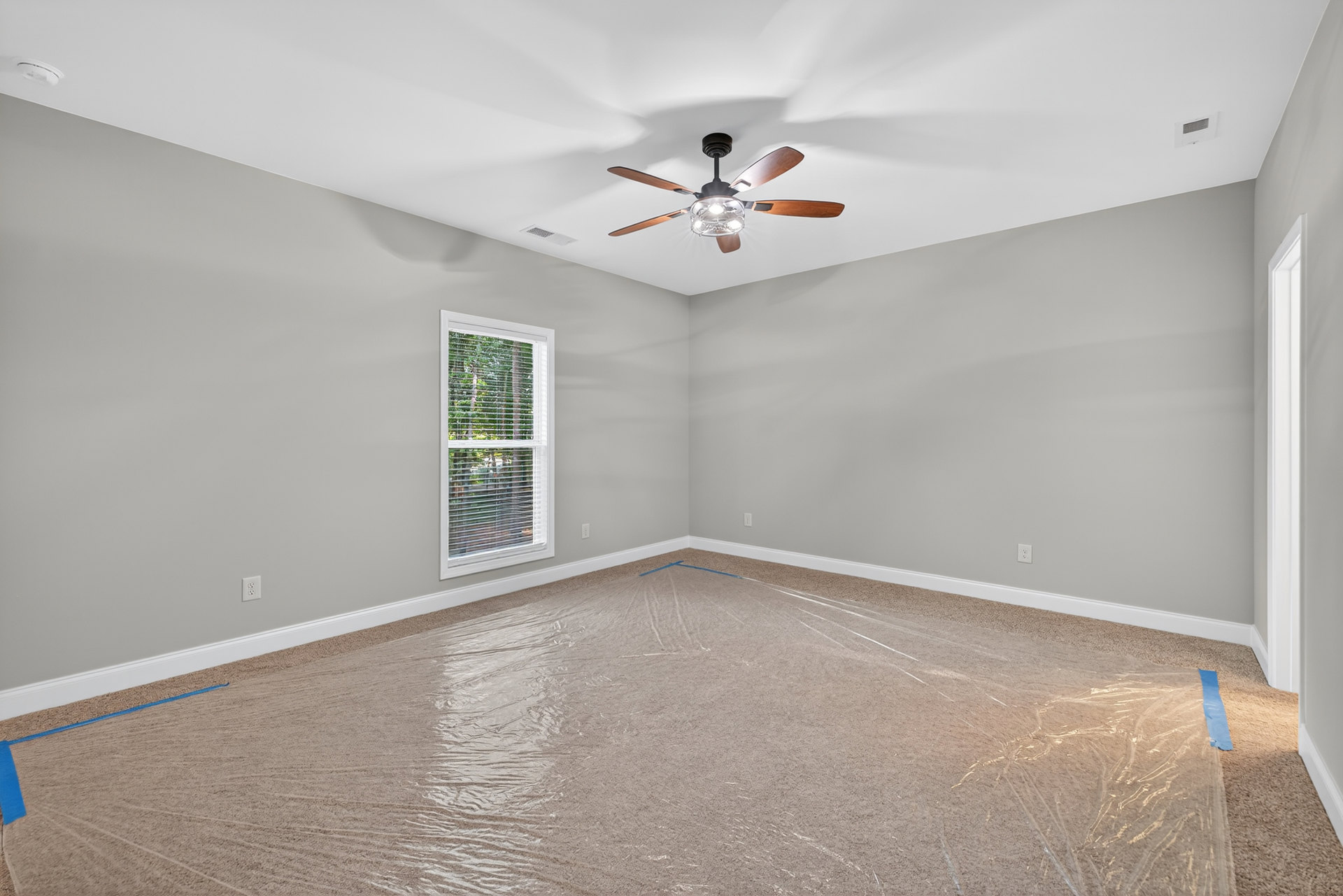 Carpeted room with ceiling fan and light fixture, window with closed blinds, plastic wrap covering part of the floor, white plaster walls and ceiling