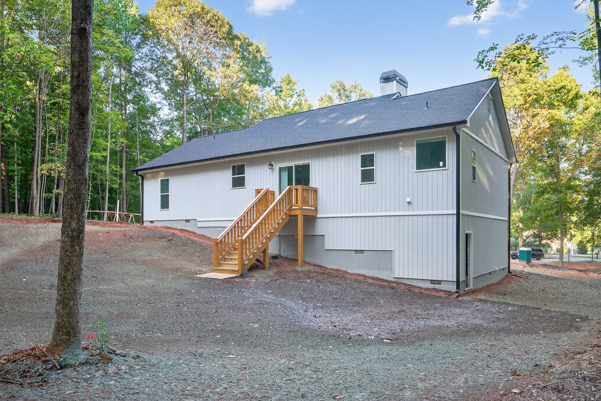 White cottage-style home with wooden exterior staircase, surrounded by garden plants and mature tree, under partly cloudy sky.