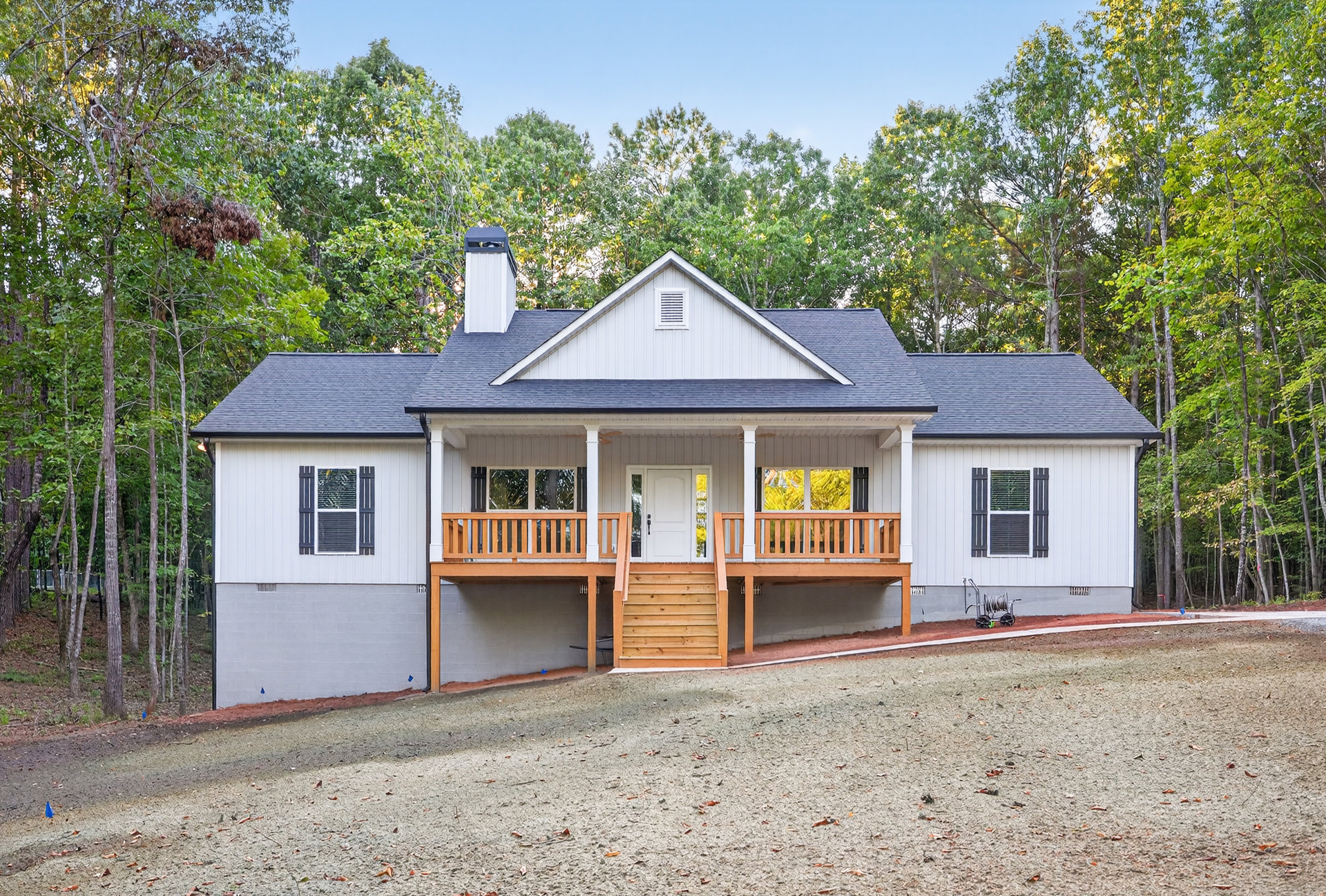 Two-story house with white siding, covered front porch, wooden stairs, gravel driveway, white-framed windows, and mature trees in the yard