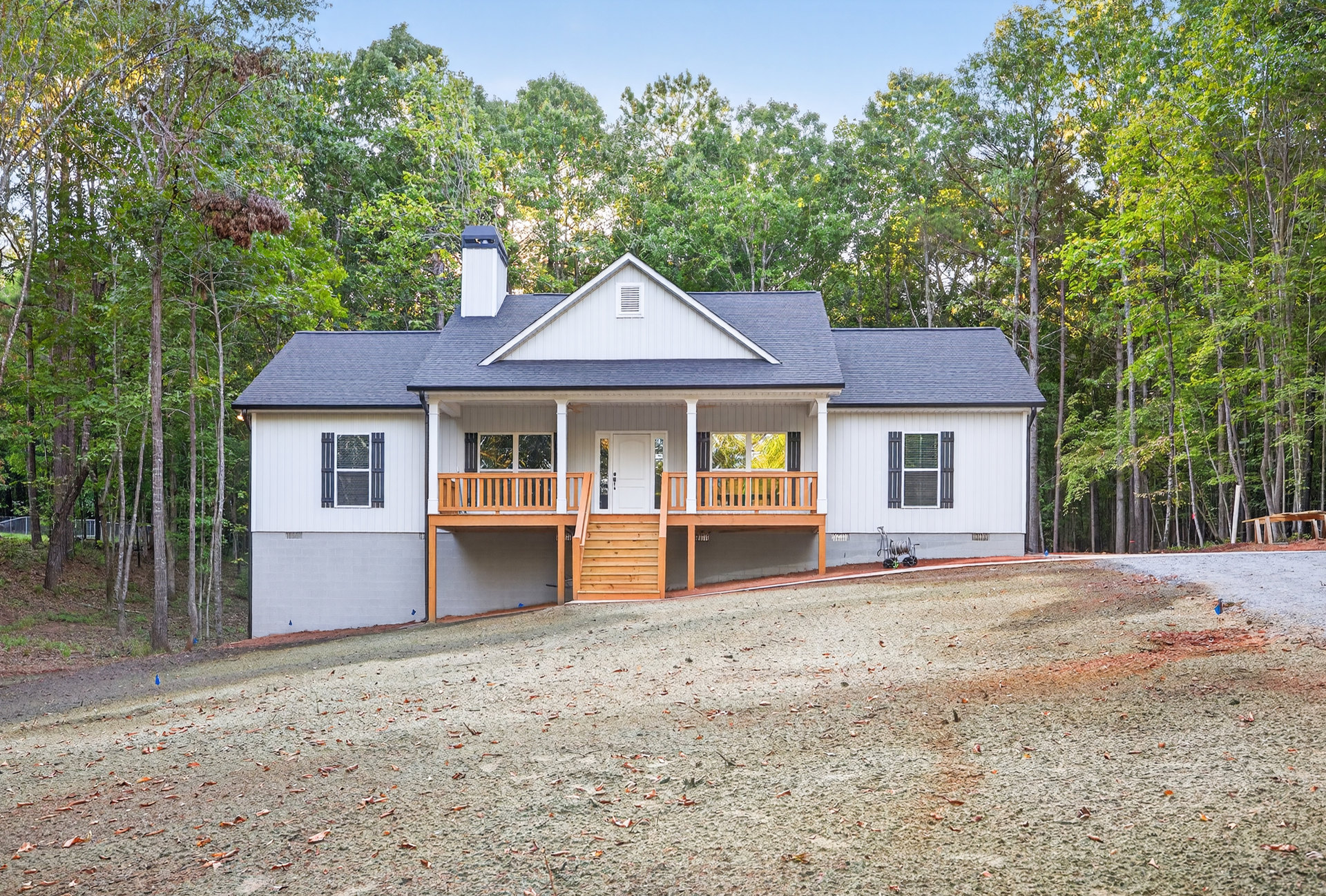 Two-story house with white siding, covered front porch, wooden steps, gravel driveway, windows with white frames and shutters, surrounded by trees and plants
