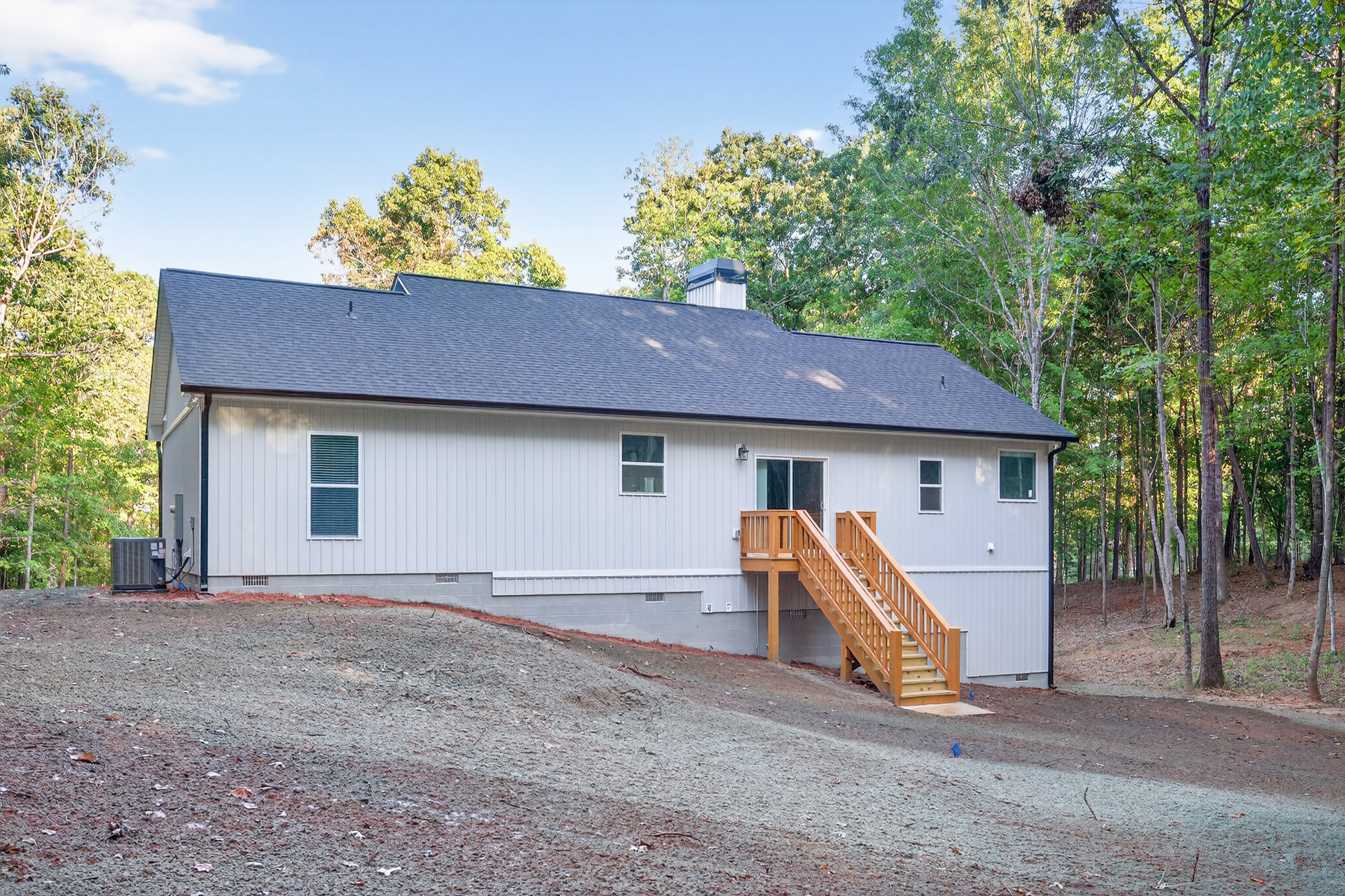 Two-story home with light siding, wooden front staircase and deck, white-framed windows, surrounded by trees and landscaping under a partly cloudy sky.