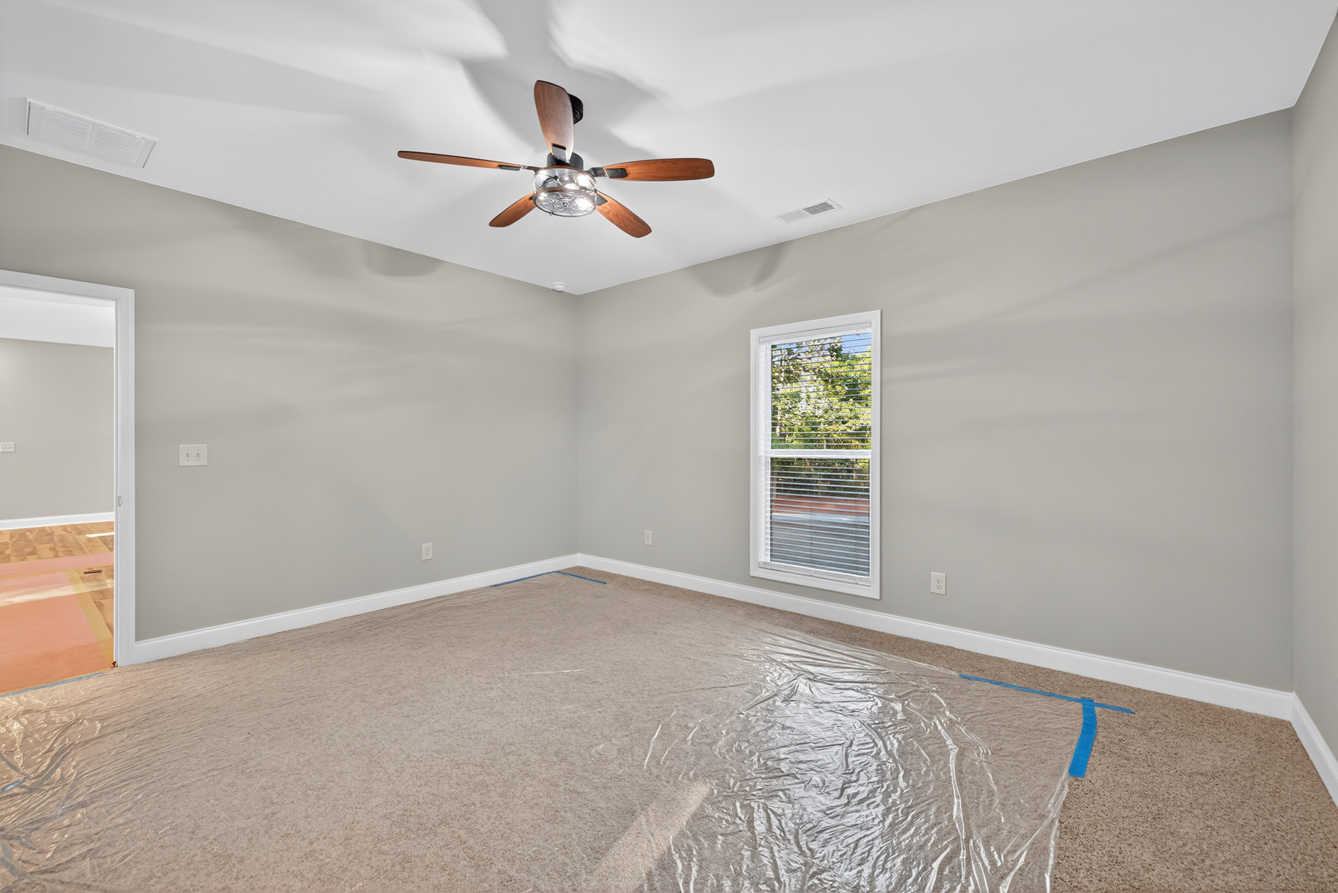 Ceiling fan with light fixture mounted on white ceiling above carpeted floor covered in protective plastic wrap, window with closed blinds on wall.