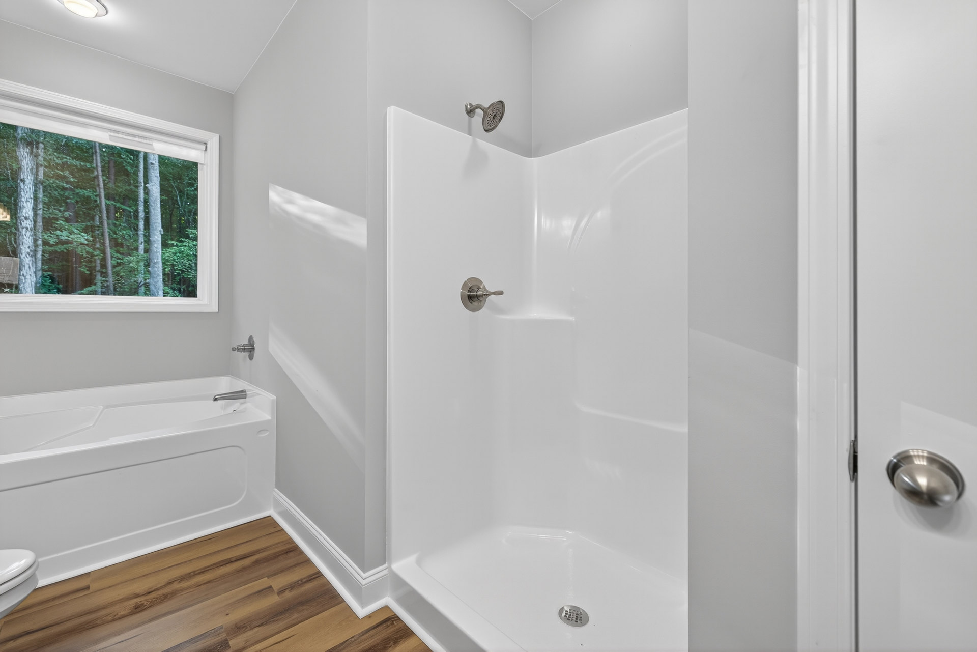 Bathroom with white tile walls, glass-enclosed shower featuring chrome handle and showerhead, freestanding bathtub near window overlooking trees, partial view of toilet seat in