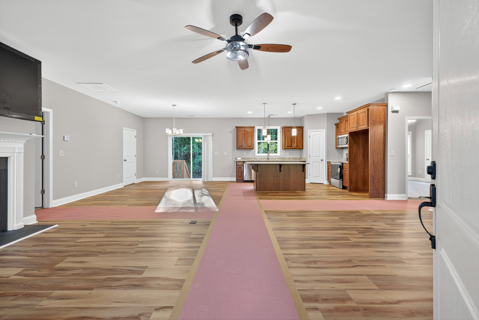 Living room with pink carpet flooring, white walls, ceiling fan with light fixture, white door with silver doorknob, refrigerator partially visible, black rectangular object