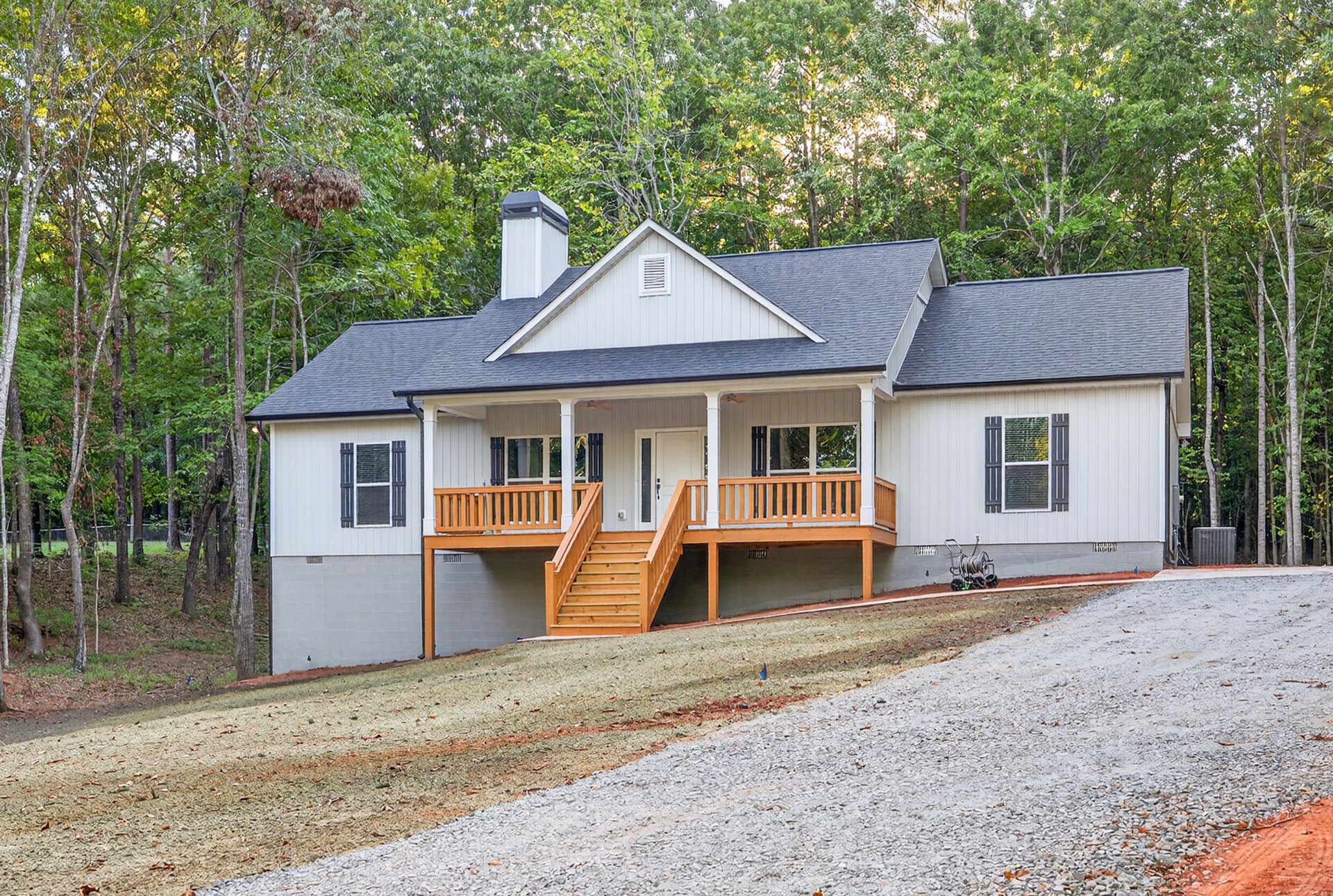 Gravel driveway bordered by mature trees, wooden staircase ascending to covered porch, white-framed windows on light-colored exterior