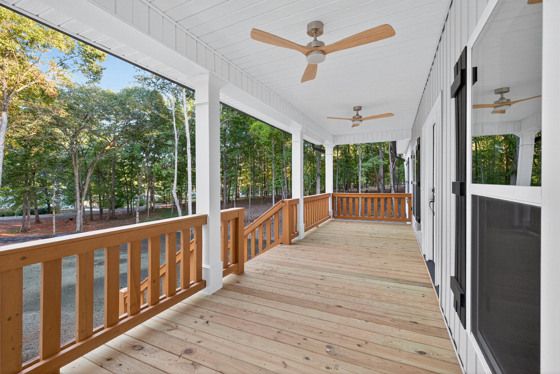 Covered porch with wood decking, ceiling fans with light fixtures, wooden railings, and trees visible beyond the fence in the background