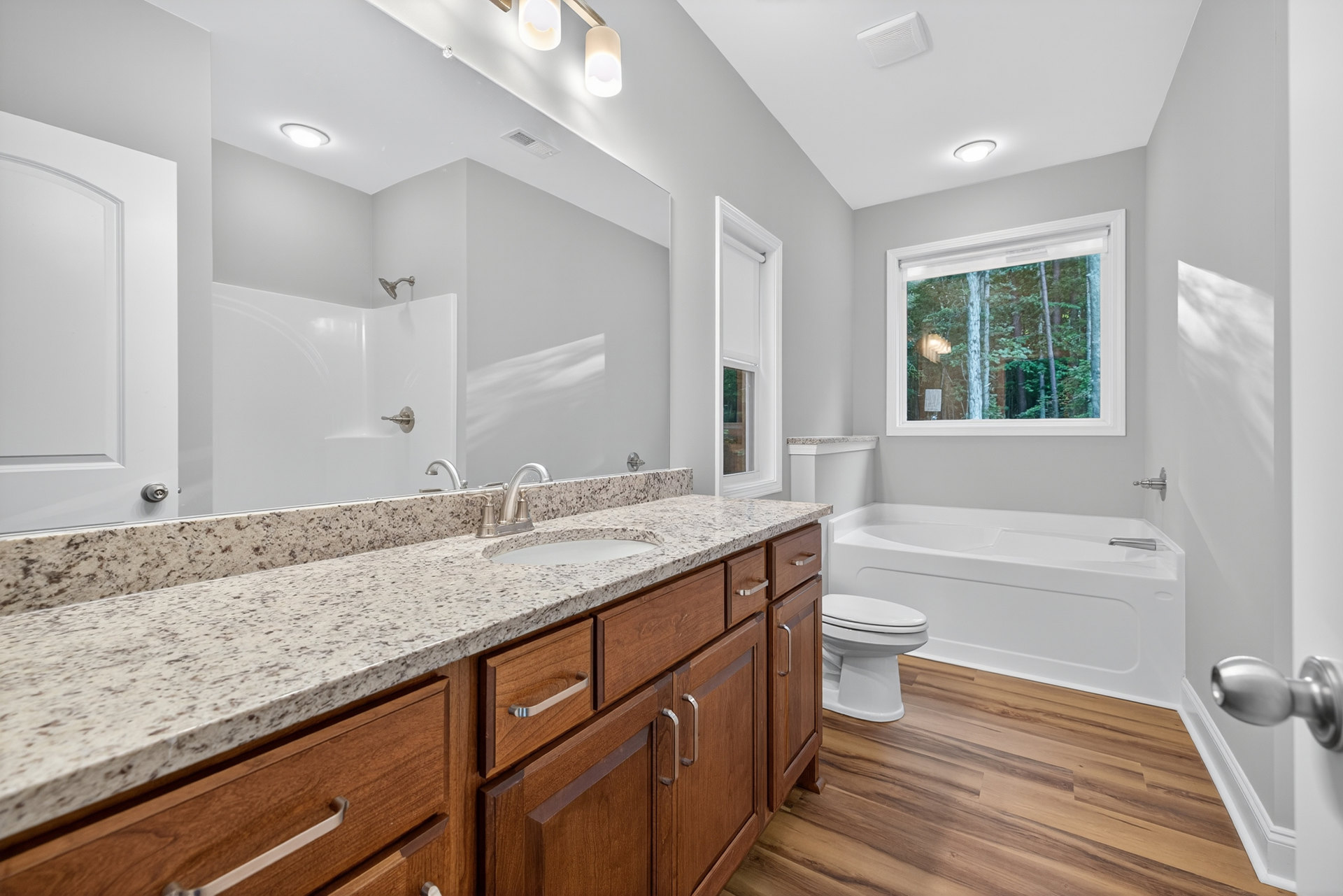 Bathroom with marble countertop, freestanding bathtub, wooden floor, white toilet, window overlooking trees, close-up of metal doorknob and light fixture
