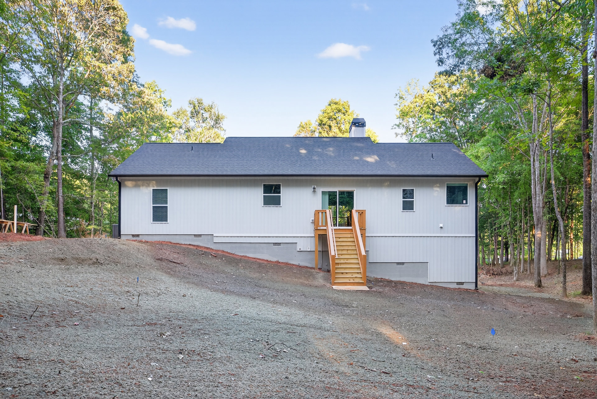 Two-story house with exterior wooden staircase, gravel driveway, white-framed window, green-framed window, blue and white trash can, surrounded by trees and plants under a cloudy