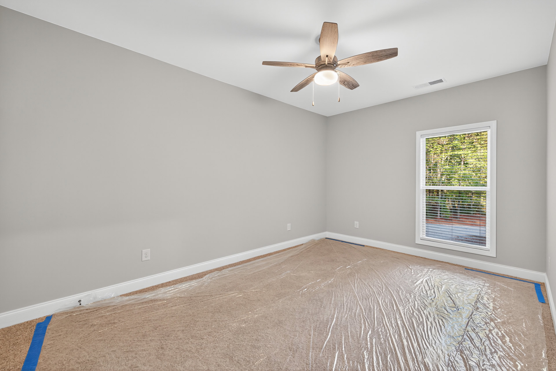 Ceiling fan with integrated light fixture mounted on white plaster ceiling, window with closed blinds, plastic wrap covering carpeted floor, close-up of wood trim and light.