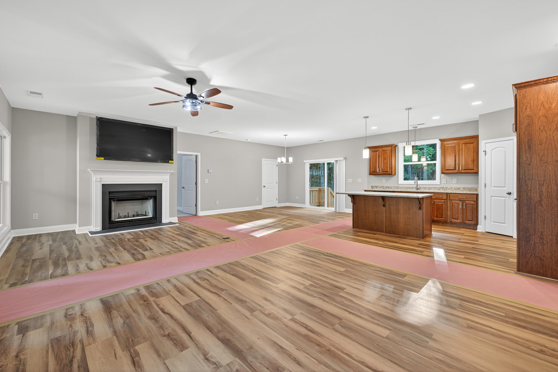 Spacious living room featuring wood flooring with pink mats, black rectangular fireplace with glass door, wall-mounted TV on a white surface, ceiling fan with light, and white