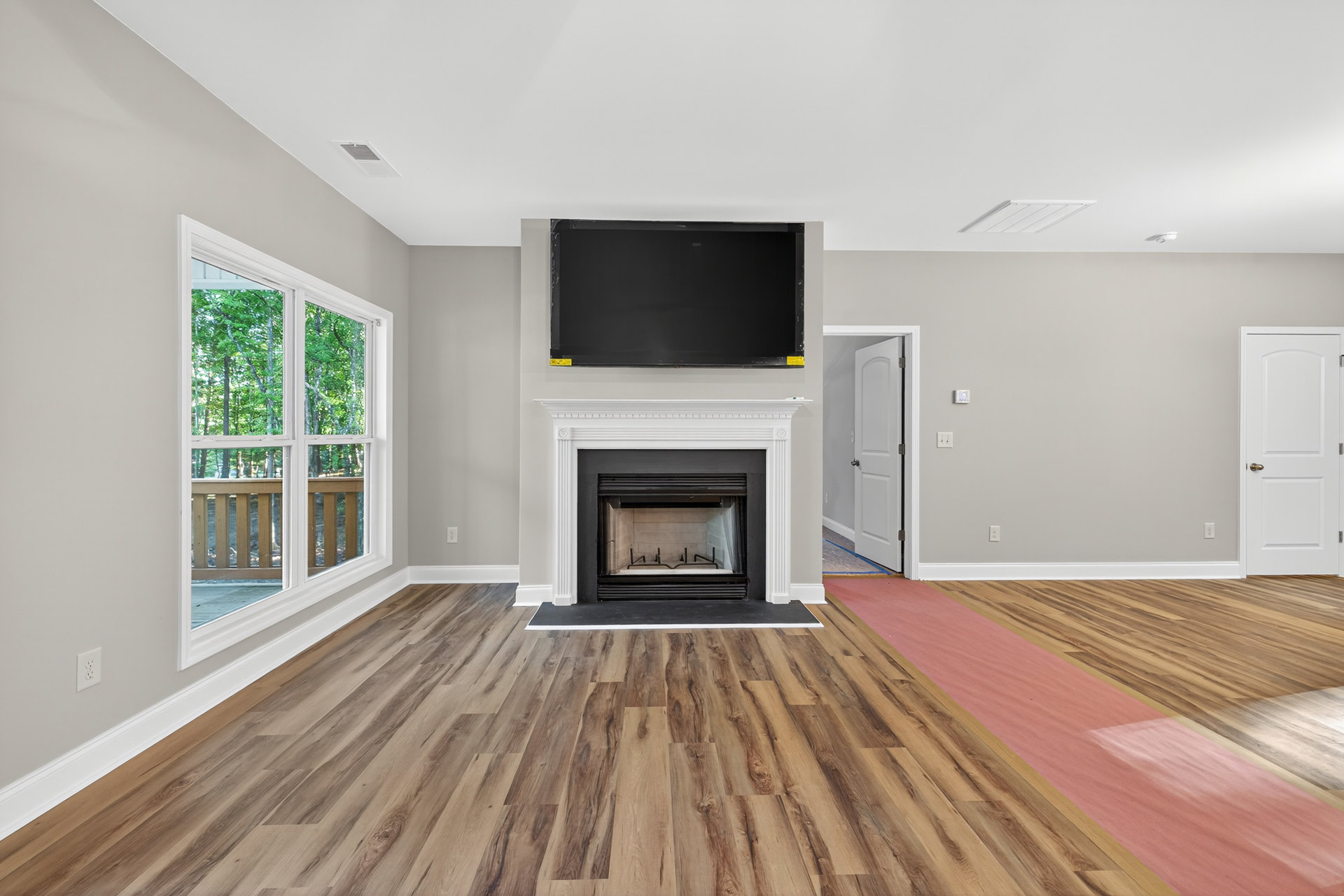 Modern living room with white walls, black-framed fireplace, hardwood floors, large window showing trees, and white door with gold handle