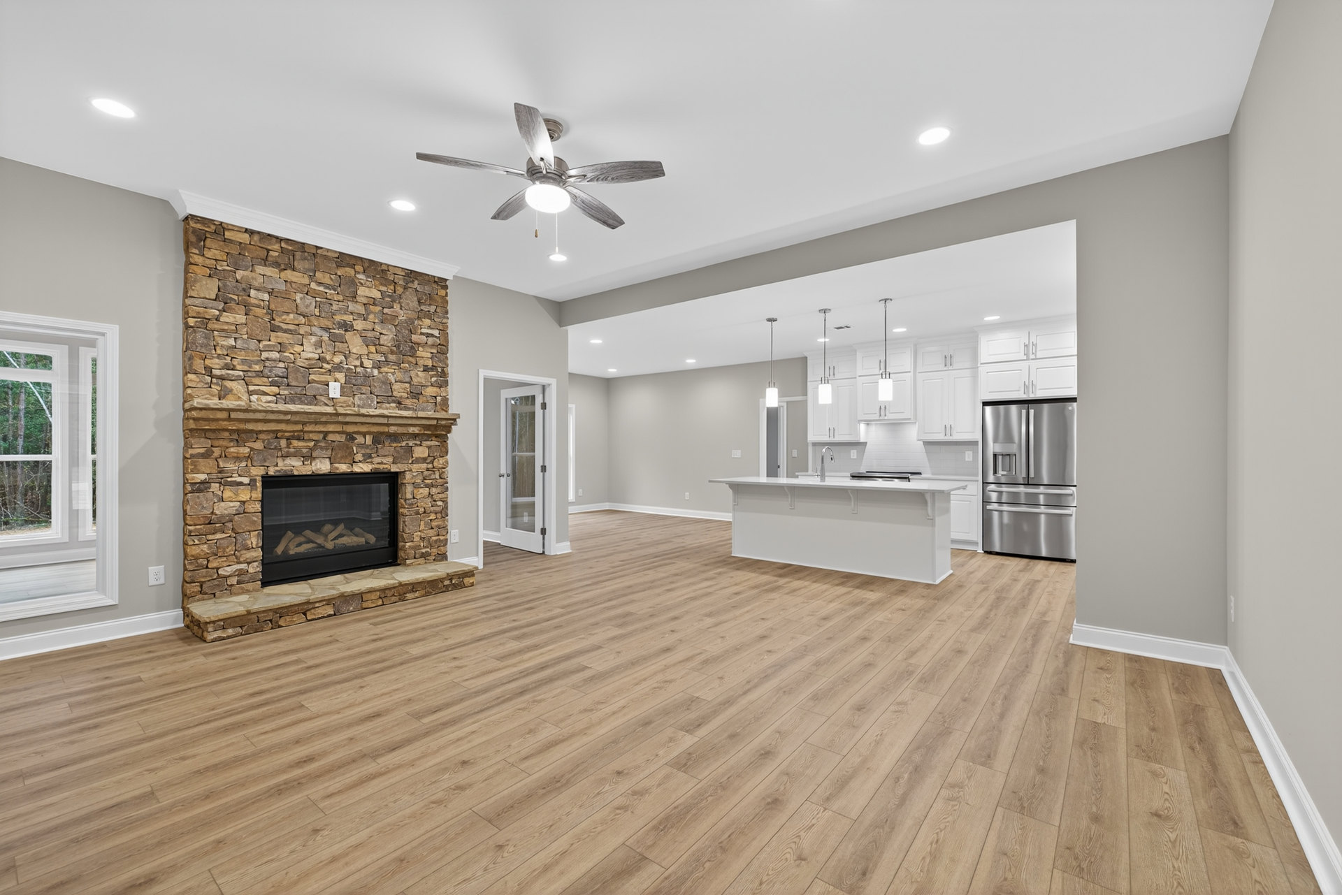 Open concept living room with stone fireplace, wood flooring, ceiling fan with light, adjacent kitchen featuring white countertops and stainless steel refrigerator